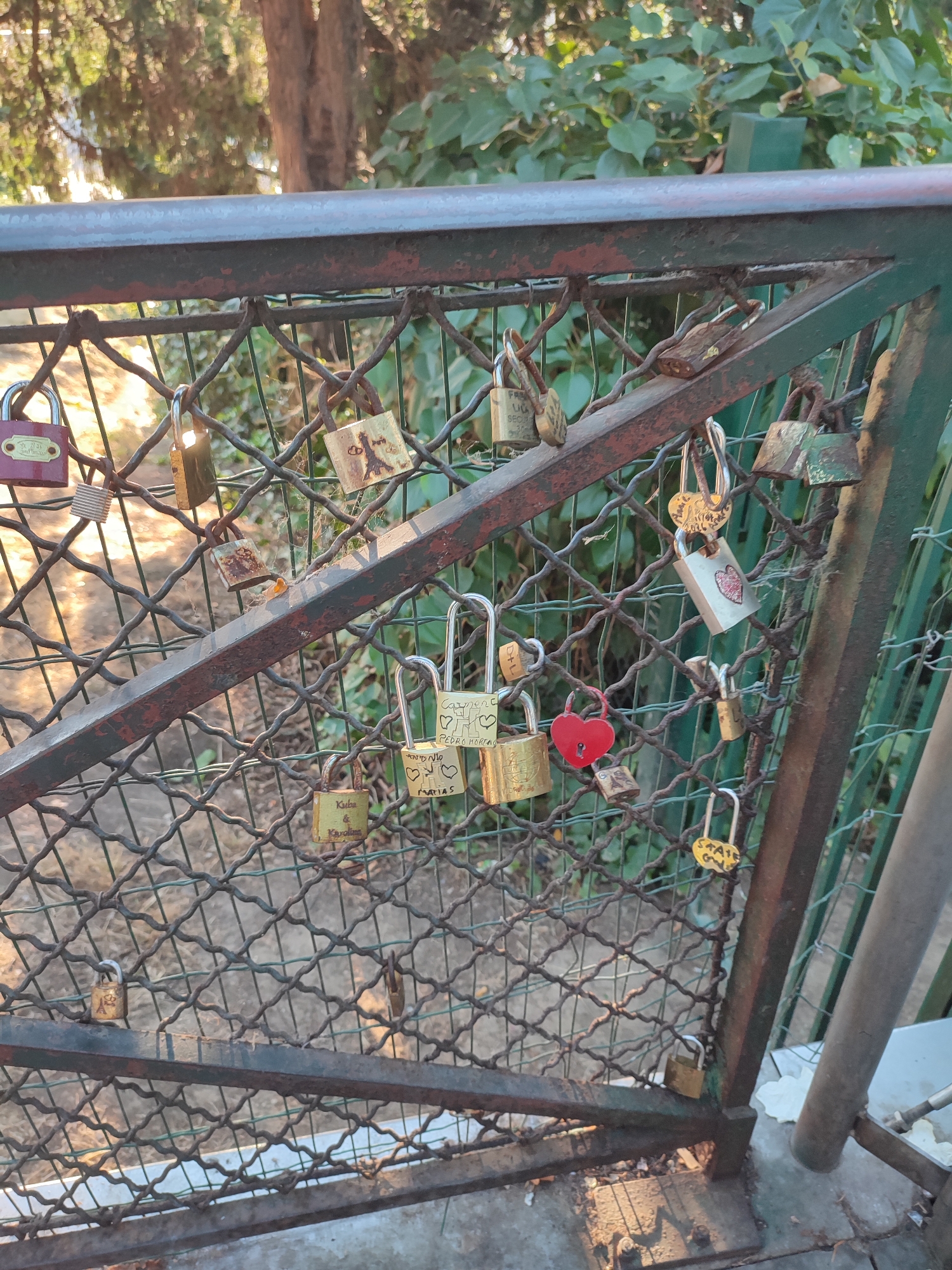 The image shows a close-up of a metal fence covered in love locks. The fence appears to be part of a bridge or walkway, with lush green foliage visible in the background. The locks are of various shapes, sizes, and colors, with some bearing inscriptions and drawings, including hearts and the Eiffel Tower. The overall scene suggests a romantic and possibly touristy location, consistent with the tradition of attaching love locks to bridges in cities like Paris, France. Sunlight filters through the trees, casting dappled shadows on the fence and locks. The ground beneath the fence is visible and appears to be a paved surface with some gravel. No people are present in the image, and the locks themselves are the sole focus.