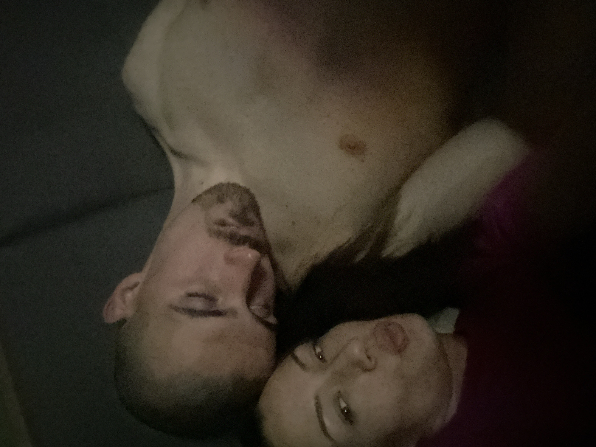 A low-angle, underexposed shot depicts two individuals. A shirtless man with a close-cropped haircut and a short beard lies on a dark surface. A woman with dark hair lies close to him, facing the camera. The location is not discernible from the image.
