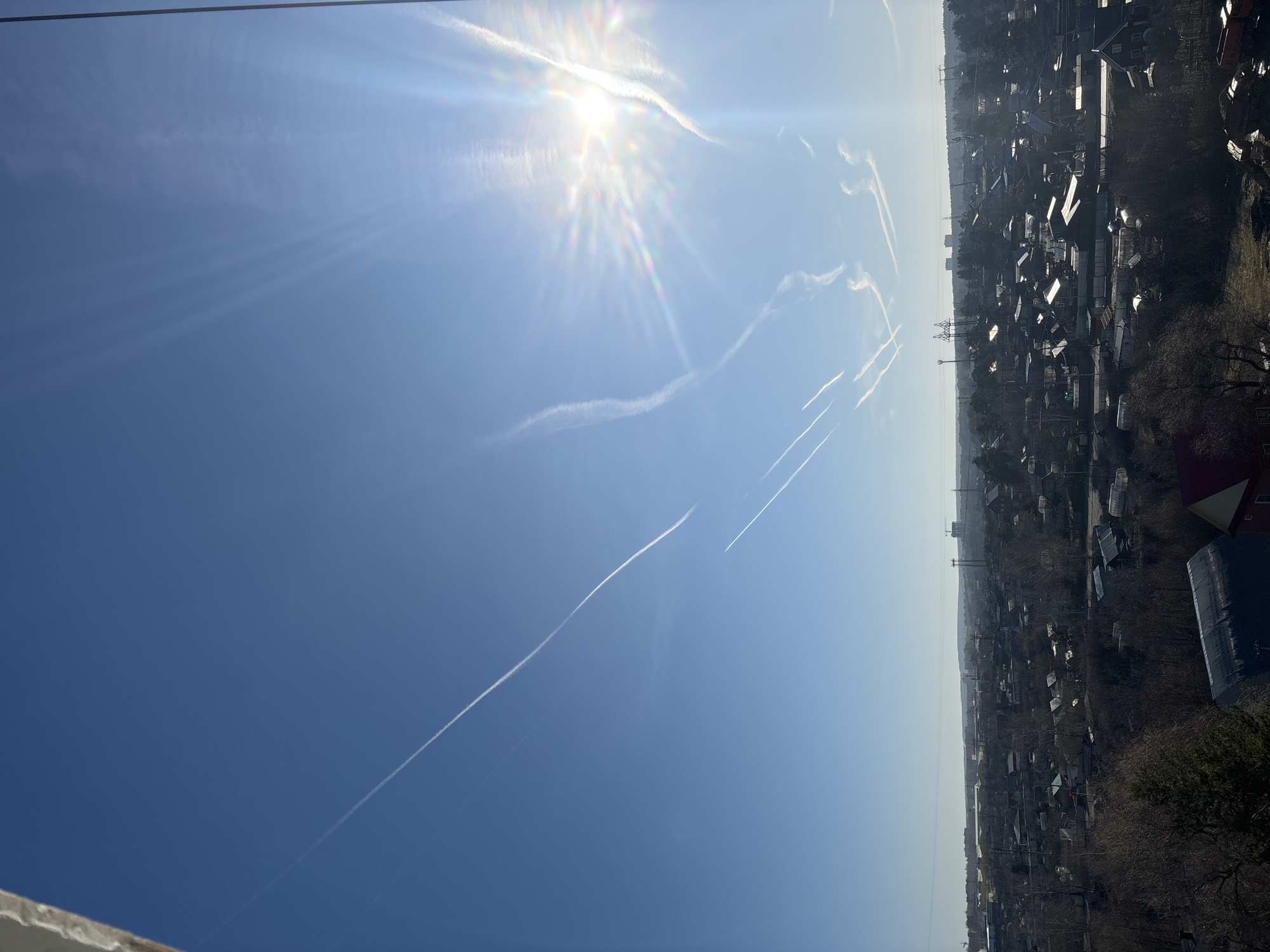 The image shows a cityscape in Chaikovsky, Russia, under a bright, clear blue sky. The sun is high in the sky, casting a strong glare. Several contrails from airplanes are visible streaking across the sky. Below, the city is composed of a dense mix of low-rise buildings, possibly residential houses, interspersed with trees. The overall impression is of a sunny day in a suburban or semi-rural area.