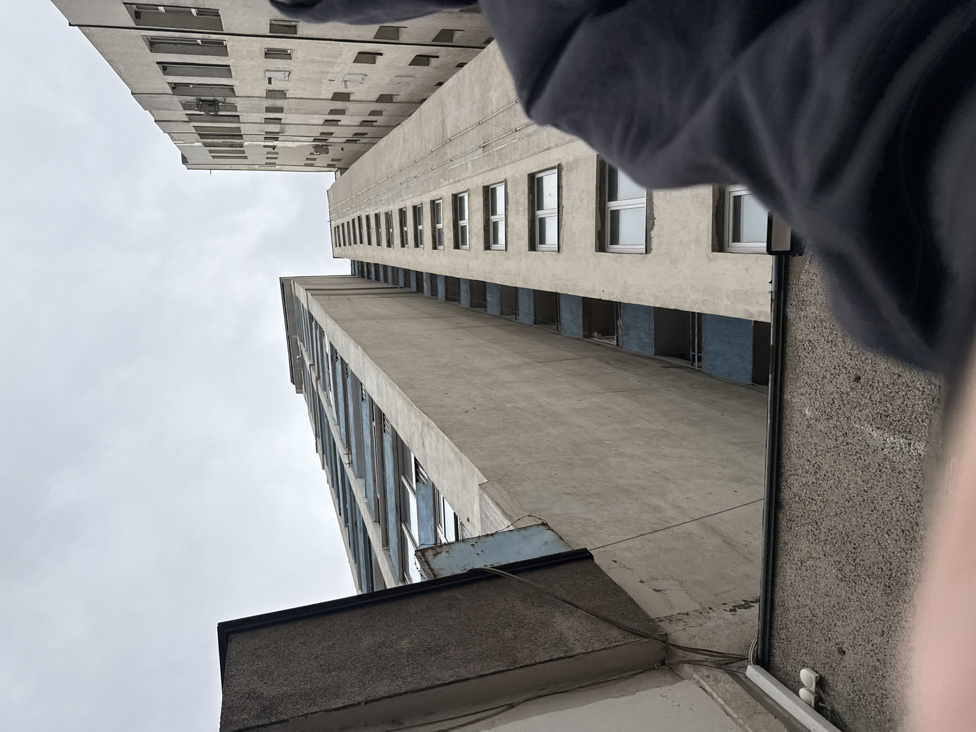 The image shows a low-angle view of two tall, concrete buildings in what appears to be an urban area. The buildings are characterized by repetitive window patterns and have a weathered, somewhat dilapidated appearance. The sky is overcast. A portion of a person's clothing or body is visible in the upper right corner of the frame. Based on the architectural style, the buildings are likely located in Sofia, Bulgaria.