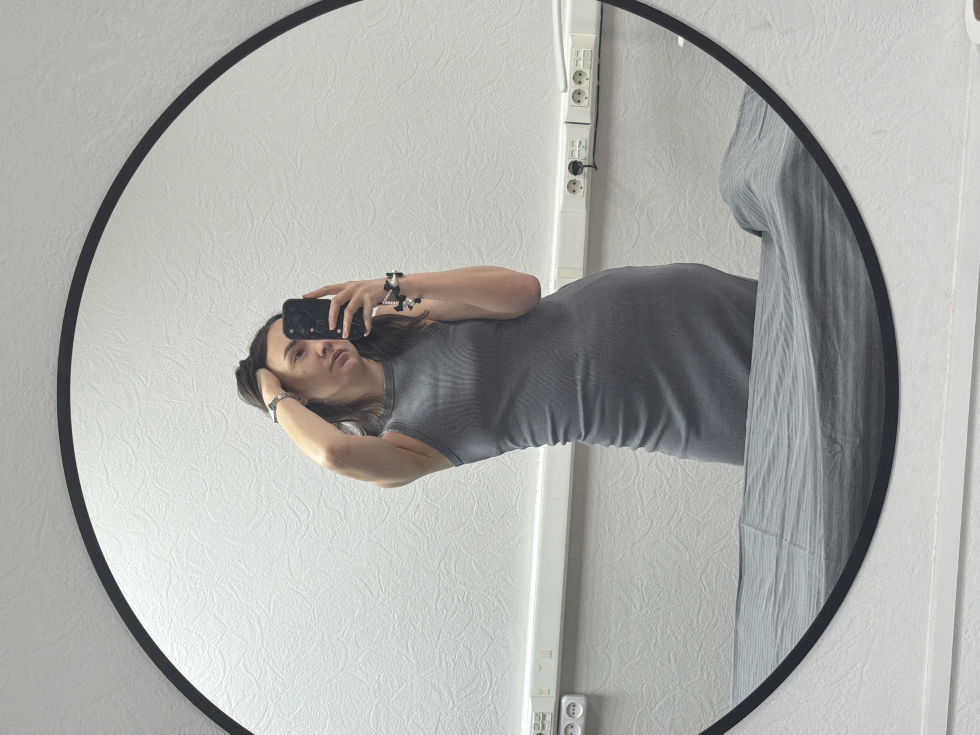 A person is reflected in a round mirror with a black frame. The person is holding a smartphone and appears to be taking a selfie. They are wearing a gray dress. To the right of the mirror, there is a vertically oriented white fixture, potentially electrical conduit, with electrical outlets and a hanging cord attached. The background is a white wall with a textured surface. The location is Chaikovsky, Russia.