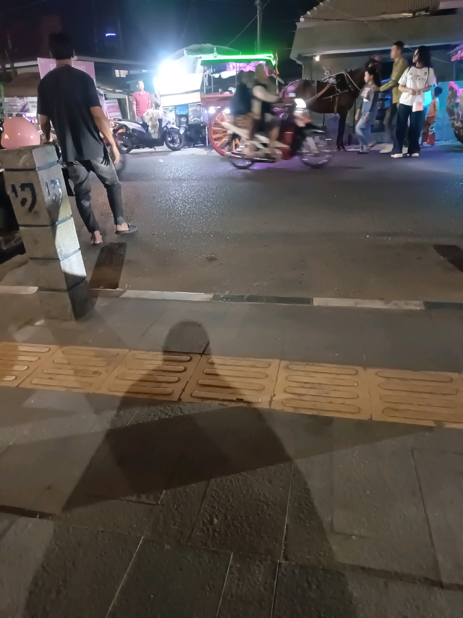 This image captures a nighttime scene on a street in Lubuklinggau, Indonesia. In the foreground, a person stands facing away from the camera, leaning against a stone pillar. The person is wearing a black t-shirt and grey pants, with black sandals on their feet. A pink helmet is visible on the stone pillar. The street surface appears wet, possibly from recent rain, and is illuminated by artificial lights from nearby stalls or businesses.
In the mid-ground, a horse-drawn carriage, likely a delman or similar local transport, is moving. Several people are visible around the carriage, including passengers and onlookers. One person, wearing a white t-shirt with "CHICAGO" printed on it, stands near the horse. Another individual in a light-colored shirt and dark pants also appears to be interacting with the scene. A motorcycle is parked or moving in the background. The environment suggests a bustling area, possibly a market or entertainment district, with various structures and bright lights contributing to the atmosphere. The overall lighting is low, characteristic of nighttime, with highlights from strong light sources.