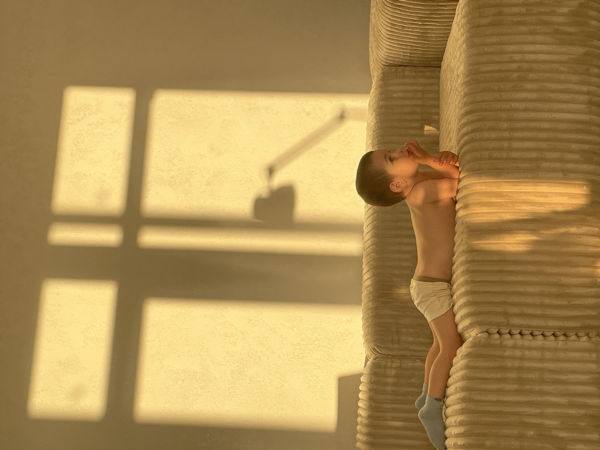 The image captures a young child lying on a ribbed sofa, bathed in warm, golden light. The child is positioned on their side, with their head resting on a cushion and their hands near their mouth. They are wearing light-colored underwear and blue socks. The sofa is a prominent object in the scene, with its textured surface creating a pattern of light and shadow.
The background is a textured wall illuminated by sunlight streaming through a window, casting distinct rectangular shadows. The play of light and shadow suggests it is either early morning or late afternoon. A faint shadow of what appears to be a lamp is also visible on the wall.
The overall mood of the image is peaceful and serene, with the warm lighting contributing to a sense of comfort and tranquility. The child appears relaxed and contemplative, possibly dozing or lost in thought. The setting is domestic, likely a living room or bedroom, with the focus on the child and the ambient light. There is no visible text in the image.