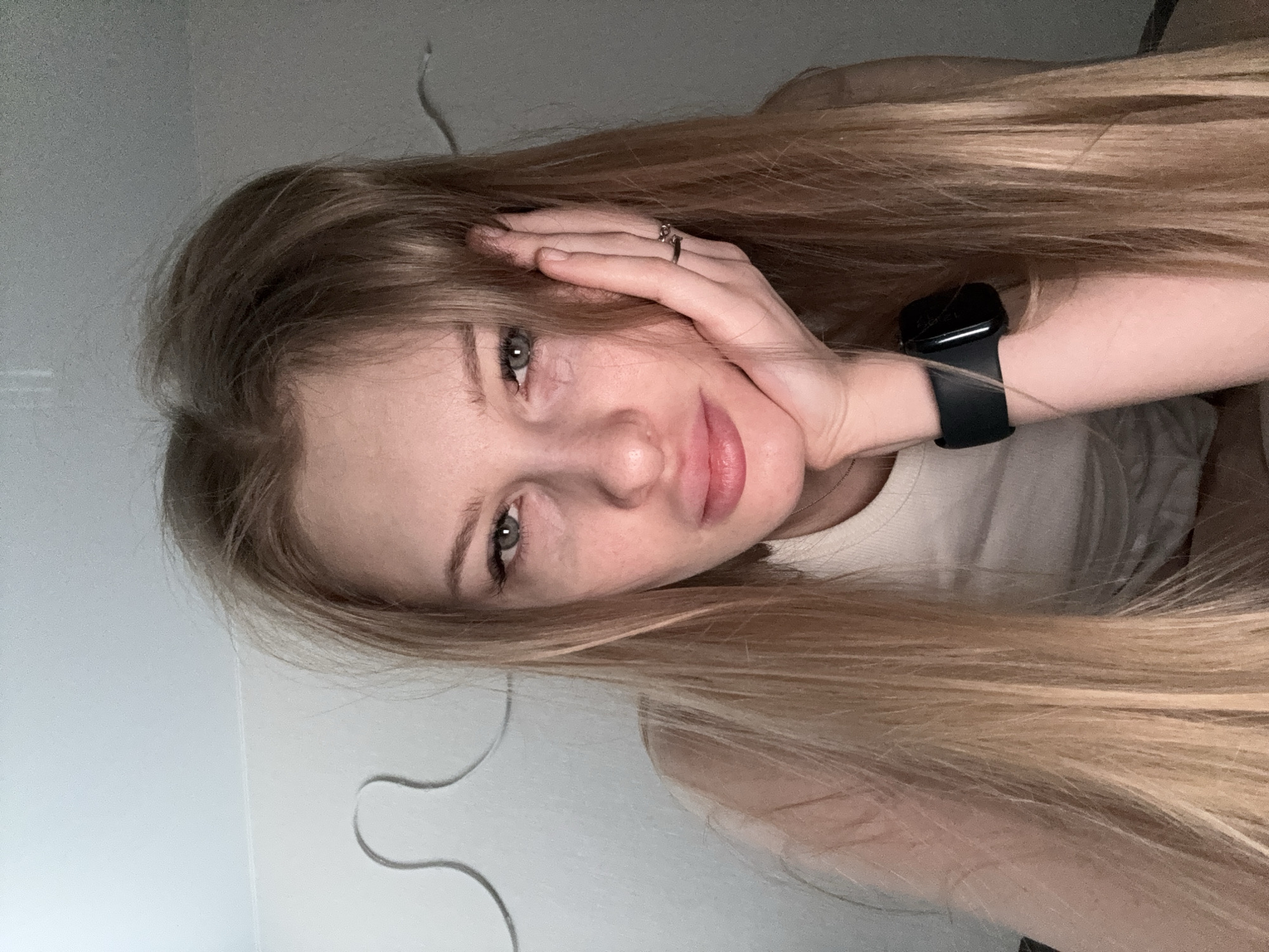 A young woman with long, straight, light blonde hair is depicted in a close-up, rotated portrait originating from Perm, Russia. Her head is gently supported by her right hand, which has a silver-colored ring on the ring finger and covers part of her forehead. She has a fair complexion with subtle freckles and soft, greenish-gray eyes that look directly at the viewer. Her lips are full and naturally pink, forming a gentle, slightly smiling expression that conveys calmness and contentment.
She is wearing a simple, light-colored top, likely white or off-white. A black smartwatch with a black band is visible on her left wrist. The background is a plain, light-colored wall, suggesting an indoor setting, possibly a room in a home. A dark, wavy line or cord is visible on the wall to the left of her head. The lighting is soft and ambient, characteristic of an interior space, potentially taken in the evening. No text is visible within the image.