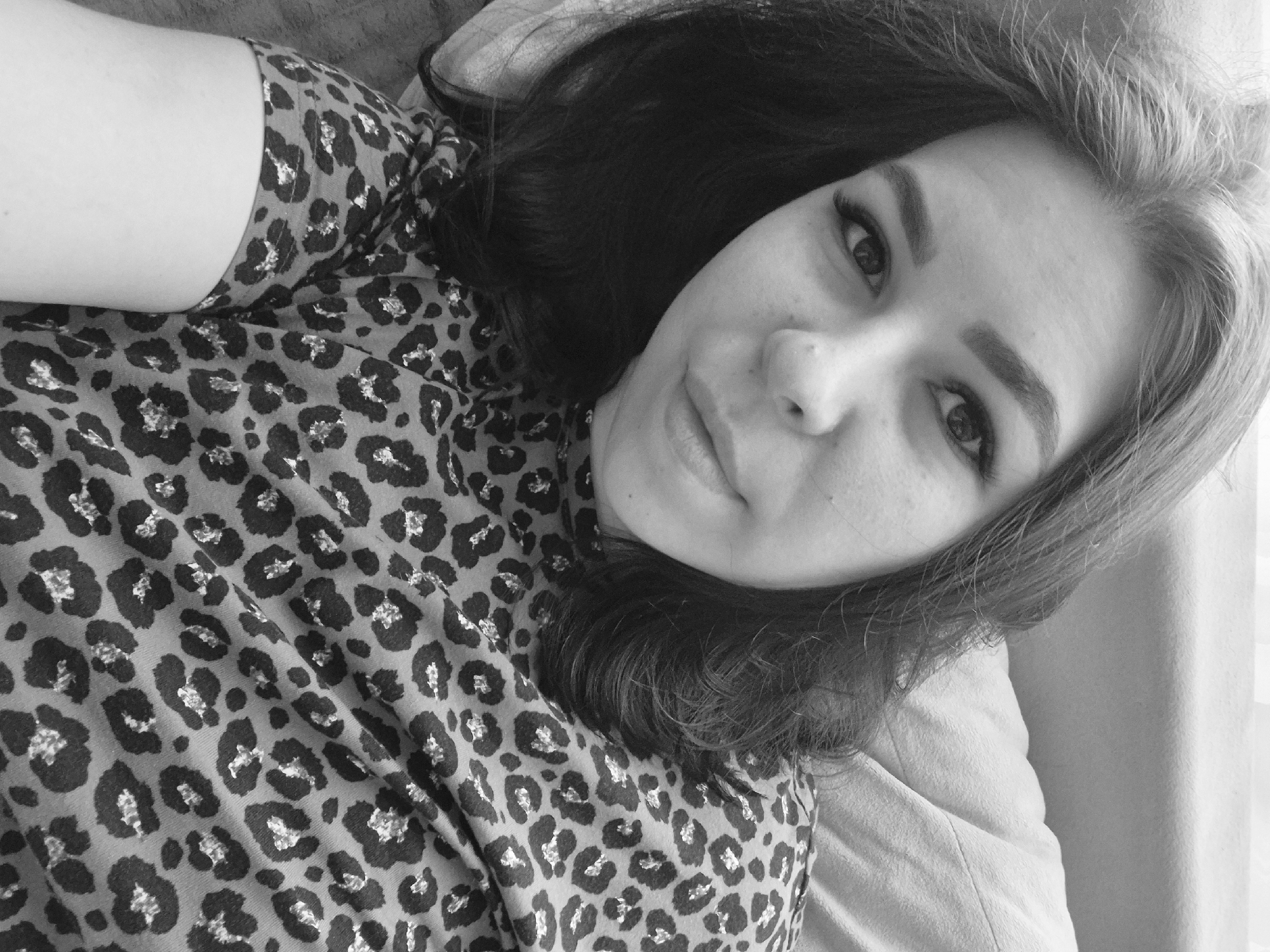 The image is a black and white selfie-style photograph of a woman with dark hair and light skin. She is lying down, angled towards the camera. She is wearing a leopard print shirt. The background is out of focus but appears to be a plain surface, possibly a bed or couch. There is no indication of the location being Tobolsk, Russia or any specific geographic location.