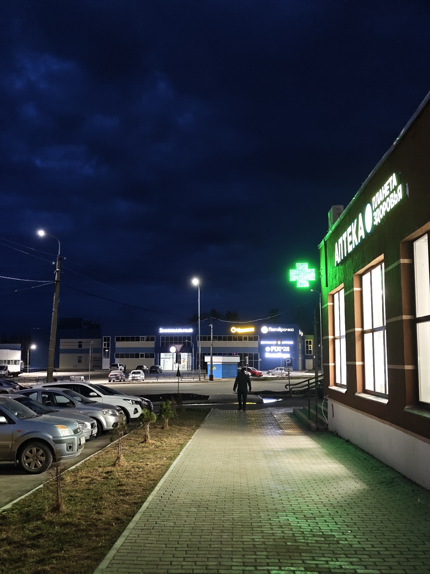 The image presents a nighttime scene in Chaikovsky, Russia. It features a pharmacy ("Аптека" in Russian) with illuminated signage and a green neon cross, indicating its function. A person is walking on the sidewalk in front of the pharmacy. Across the street, other commercial buildings with neon signs are visible, including what appears to be a "Пятёрочка" (Pyaterochka) supermarket. A line of parked cars is visible on the left. The sky is dark, indicating that it is night, and streetlights provide additional illumination. The sidewalk is paved with bricks. There are some young trees next to the cars.
