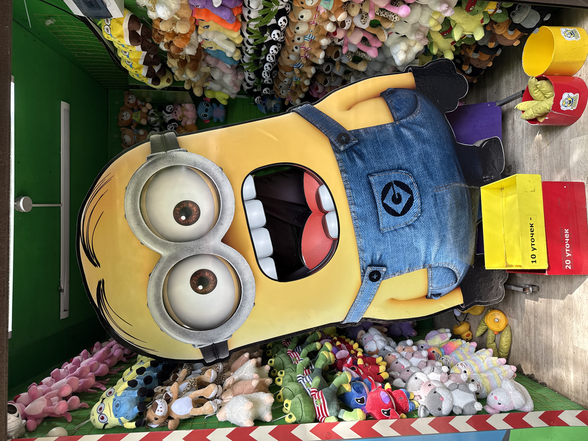 The image depicts a fair or carnival game in Izhevsk, Russia. The game involves throwing objects into the open mouth of a large minion figure. The minion is wearing denim overalls and has two large goggles with brown eyes. The mouth is open, revealing teeth and a red interior.
The background is filled with stuffed animals, presumably the prizes for the game. There are pink, blue, brown, panda, and other colored plush toys stacked above and below the minion.
To the right of the minion are various colored buckets and containers, some containing what appear to be green plush toys. The sides of the booth are marked with the text "10 уточек" and "20 уточек" in Russian, indicating the number of ducks or targets involved in the game. A red and white striped barrier runs along the bottom edge of the scene. The game appears to be indoors.
