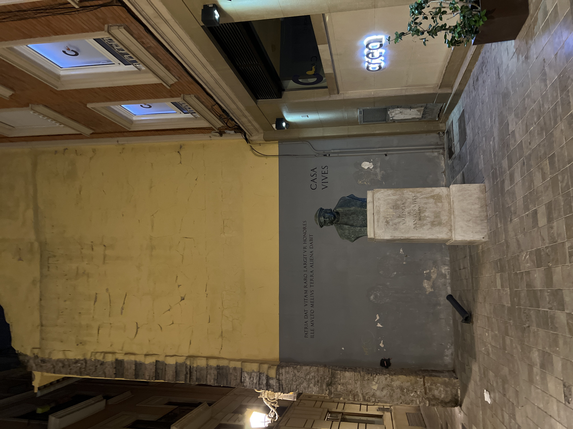 The image shows a narrow alley in València, Spain, featuring a monument dedicated to Juan Luis Vives. The monument includes a bronze bust mounted on a stone plinth. The inscription reads: "PATRIA DAT VITAM RARO LARGIT VR HONORES ILLE MVLTO MELIVS TERRA ALIENA DABIT." and under the bust, "CASA VIVES VALENCIA JUAN LUIS VIVES MCMLXVI." One wall is painted yellow with visible signs of aging, while the other is gray, with the monument placed against it. The alleyway floor is paved with light-colored bricks. Overhead, there are architectural details, including windows with visible light, recessed lighting, and signage reading "area." The alley is a mix of old and new, with modern elements interspersed with historical architectural details.