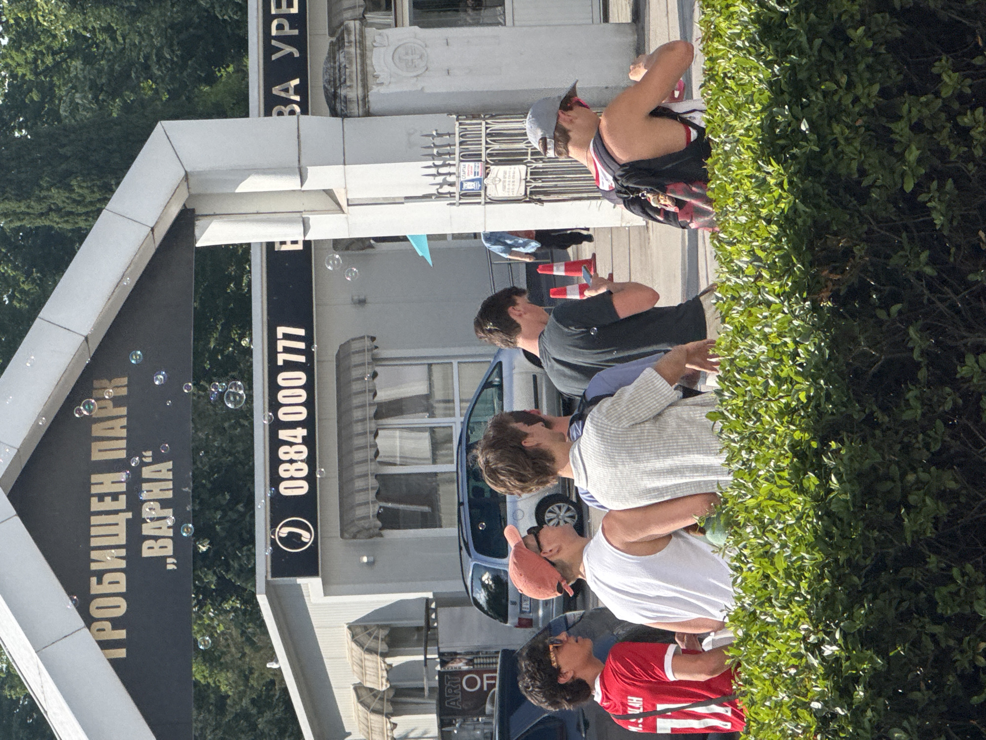 The image captures a sunny outdoor scene at the entrance of a park or cemetery in Varna, Bulgaria. A prominent black sign with gold Cyrillic lettering reads "ТРОБИЩЕН ПАРК" (likely a misspelling of "Гробищен Парк" - Cemetery Park) and "„ВАРНА“", situated above a white, angular archway structure. Numerous soap bubbles are actively floating in the air around the entrance.
In the foreground, a lush green hedge partially obscures a group of approximately six young adult males walking away from the viewer. From left to right, key individuals include: a male wearing a red sports jersey with "M. SALAH 11" on the back and sunglasses; a male in a white tank top and an orange baseball cap with sunglasses; a male in a light-colored striped shirt; a male in a black t-shirt looking down at a mobile phone; and a shirtless male wearing a grey baseball cap, red shorts, and a black backpack.
Behind the group and the archway, a white building with multiple windows and striped awnings is visible. A smaller black sign on this building displays a phone icon and the number "0884 000 777". To the right, part of another sign reads "ВА УРЕ". A metal gate with decorative spikes and an unreadable smaller sign is positioned between two white stone pillars. A silver minivan is parked in the mid-ground, and two orange traffic cones are placed near the gate. Tall green trees form a dense background, suggesting a park-like environment.