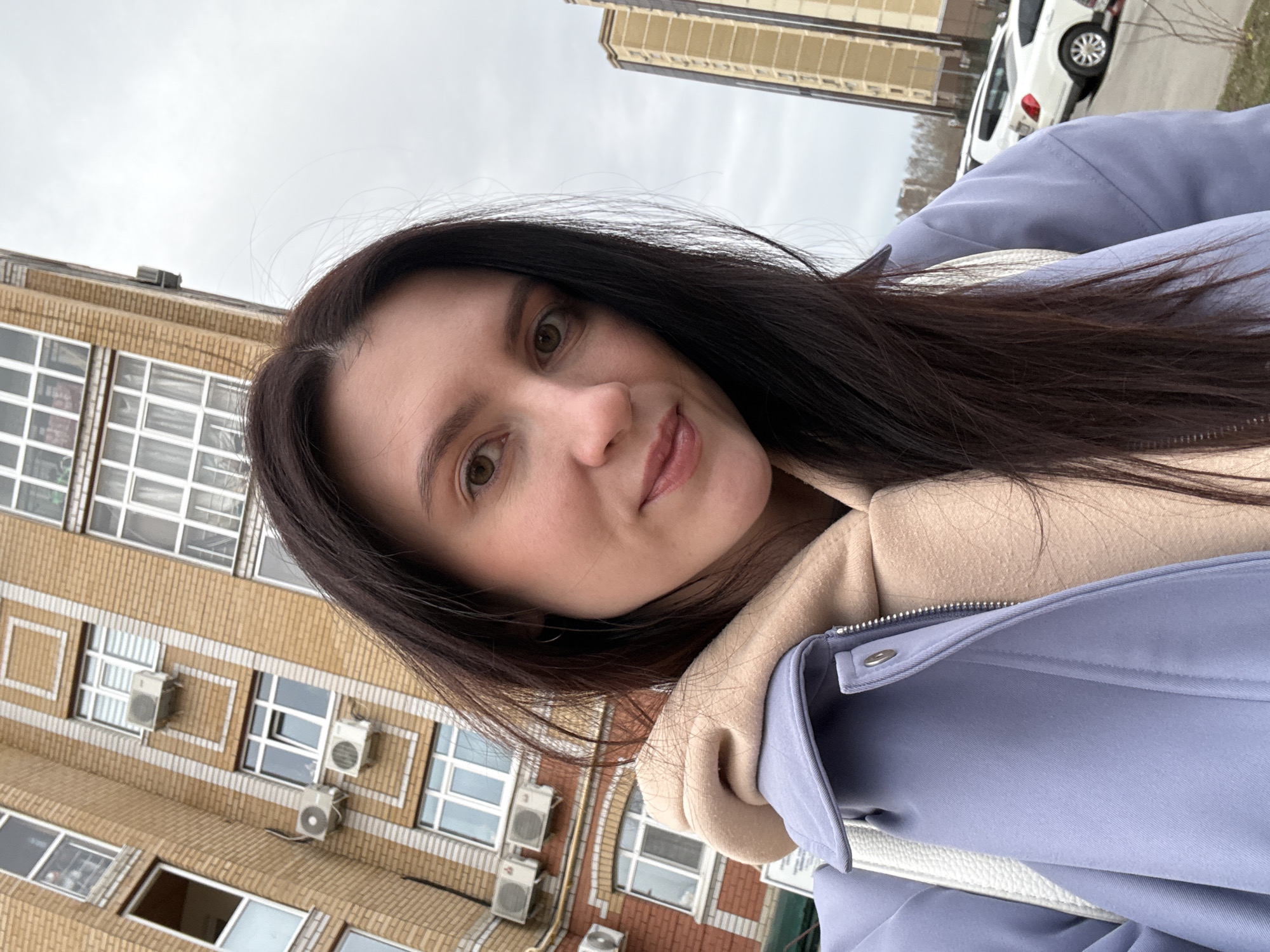 A woman with long, dark brown hair and fair skin is posing for a selfie outdoors. She's wearing a light blue jacket over a tan-colored hooded sweatshirt or sweater. The building behind her is a multi-story brick building with rows of windows and multiple air conditioning units mounted on the exterior. The sky is overcast and gray. In the background, there is a white car and another taller building. I cannot confirm the location in the city of Kazan, Russia.