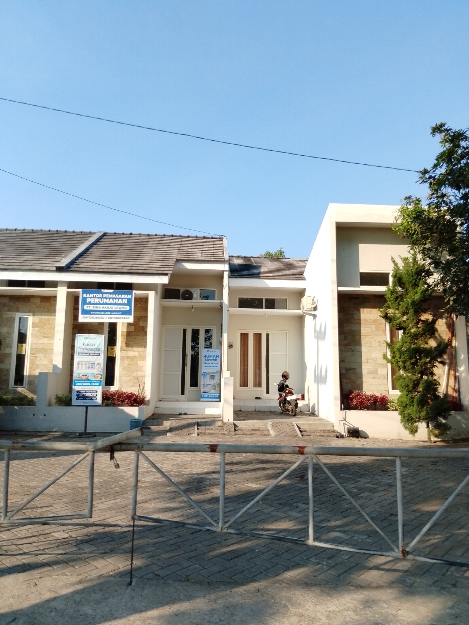 The image captures a sunny day in the city of Pasuruan, Indonesia, specifically in front of a housing complex. The main focus is a row of modern houses with white and beige facades, featuring tiled roofs. On the left, a sign indicates "KANTOR PEMASARAN PERUMAHAN" (Housing Marketing Office) for "PT. BINA HARJO UTOMO", with contact information. Another sign with "BINA LAND Project" is also visible. To the right, a similar house has a sign that says "RUMAH MEWAH" (Luxury House). A person is seated on a red motorcycle parked on the steps leading to one of the houses. The area is paved with interlocking bricks, and a metal barrier gate, partially open and locked with a padlock, stands in the foreground. A tall, green coniferous tree stands to the far right, and power lines are visible against the clear blue sky.