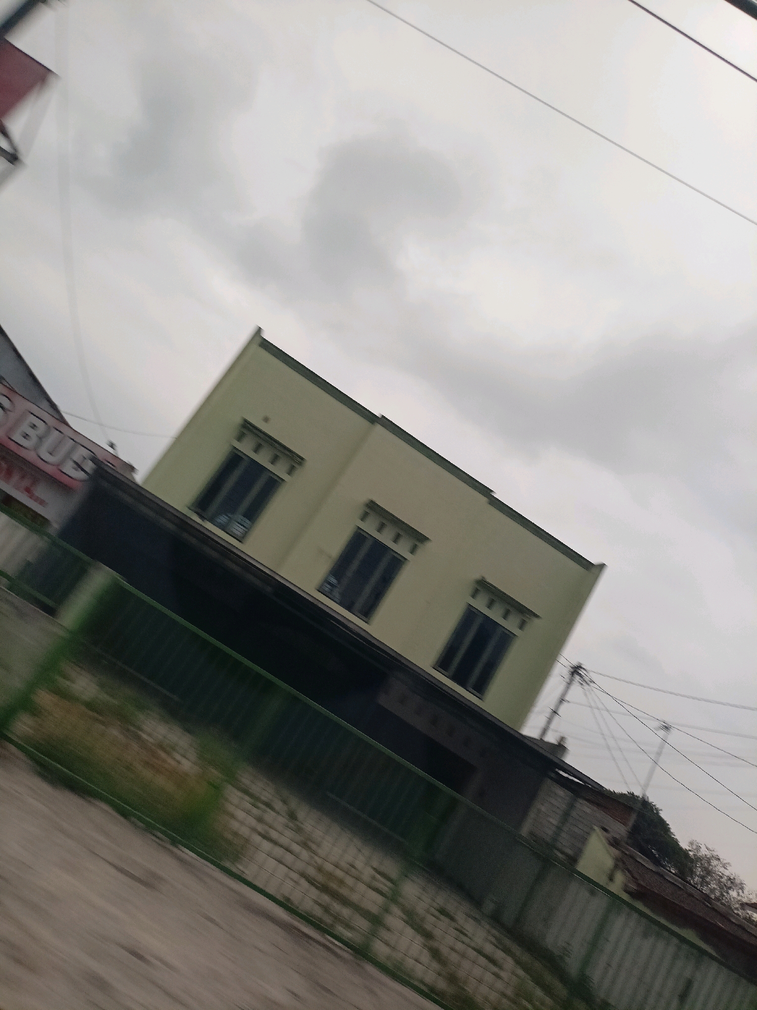 The image is a tilted, blurry photograph of a light green building with three dark-paned windows on its upper level. The building has a darker colored roof overhang. To the left of the green building, another building is partially visible with red lettering that appears to say "BUS". A green fence runs across the foreground of the image, and beyond it, there is a grassy area with some debris. The sky is overcast with clouds. The scene appears to be in an outdoor, possibly urban or suburban environment. The image is taken from a moving perspective, indicated by the motion blur.