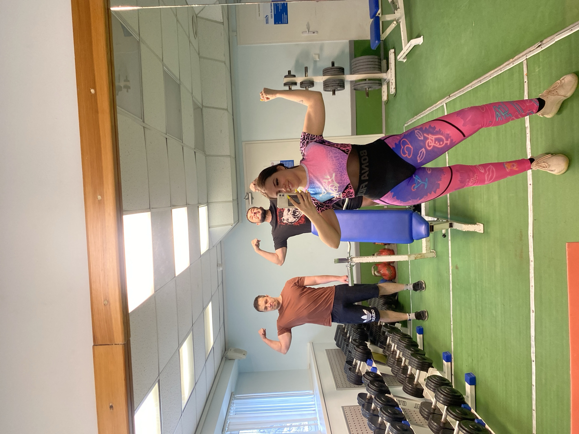 Three people are posing in a gym. A woman is taking a selfie in the mirror with two men behind her. All three are flexing their biceps. The gym has weightlifting equipment, including dumbbells, barbells, and a weight bench. The gym has a green floor, a white ceiling with fluorescent lights, and white walls. The location is Chaikovsky, Russia.