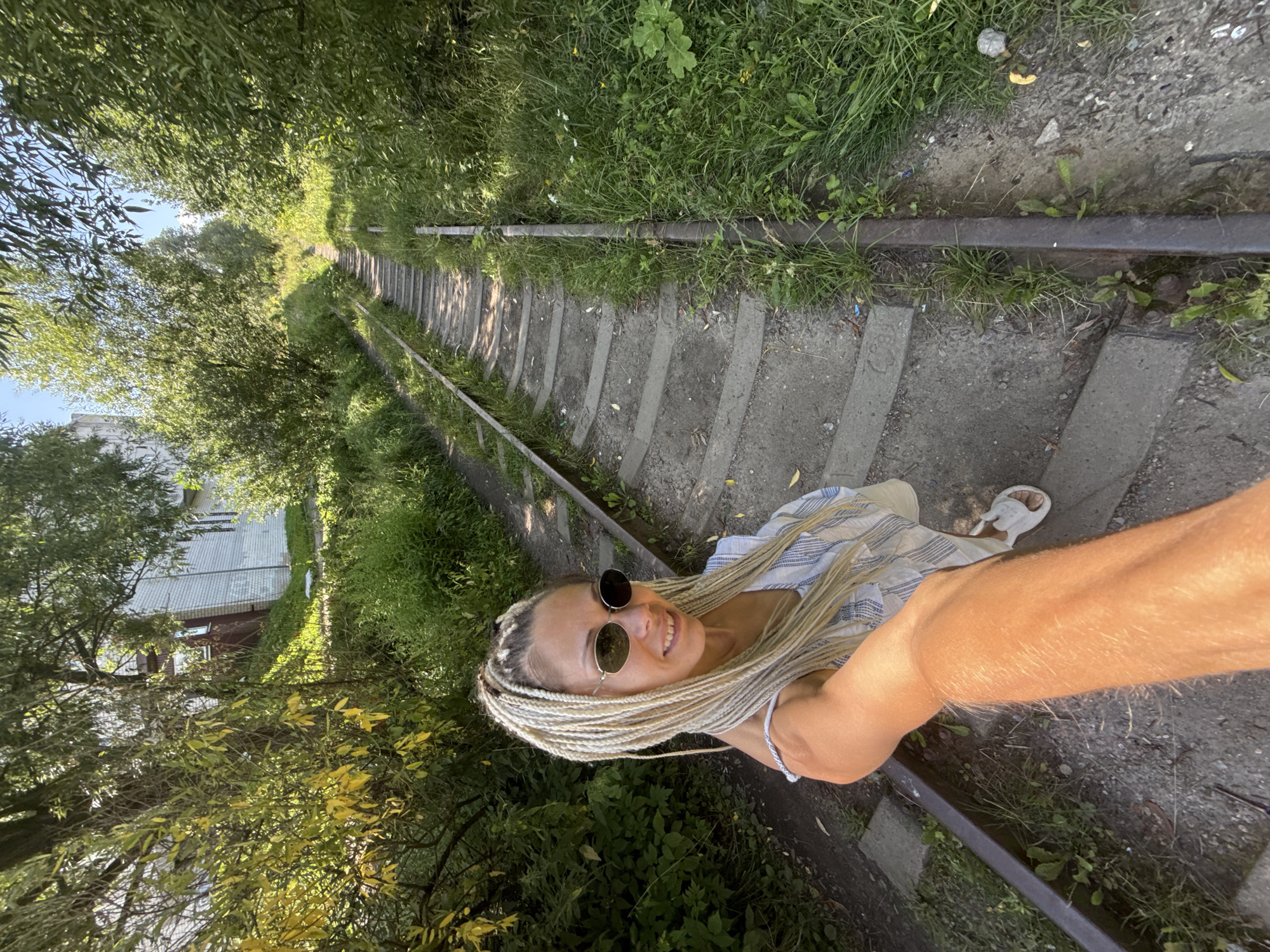 A woman with blonde braided hair and sunglasses takes a selfie outdoors. She is standing on a set of old railway tracks that are overgrown with grass and weeds. In the background, there is a white building with a sloped roof, surrounded by trees. The image appears to be taken on a sunny day. The location is the city of Rusanovo, Russia.