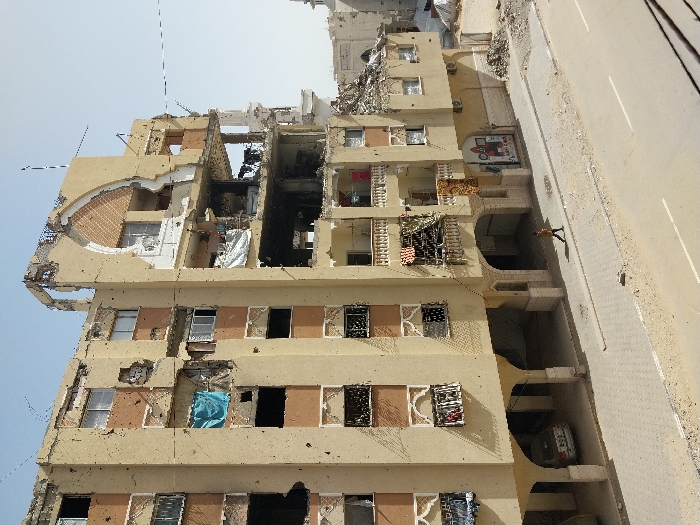 The image displays a severely damaged multi-story building in Khan Yunis, Palestinian Territory, under a clear, light blue sky. The tan-colored structure exhibits extensive structural compromise, including large sections of missing walls, exposed interior rooms, and shattered windows across its visible seven to eight floors. Significant damage is evident on the upper levels, where large portions of the facade and roofline are absent, revealing internal brickwork and rebar. Various pieces of fabric, including a prominent blue cloth, are visible hanging from damaged windows and balconies.
At ground level, a paved sidewalk runs parallel to the building, bordering an asphalt road with a faded white center line. A lone individual, a man in a dark shirt and light trousers, walks from left to right on the sidewalk. Below the building's arched ground-floor supports, a silver sedan is parked in an open covered area. To the right, adjacent structures, including a partial view of what appears to be a mosque with a distinct minaret-like element, also show signs of significant damage and accumulated debris. The immediate environment includes patches of dirt and rubble alongside the road and sidewalk.
