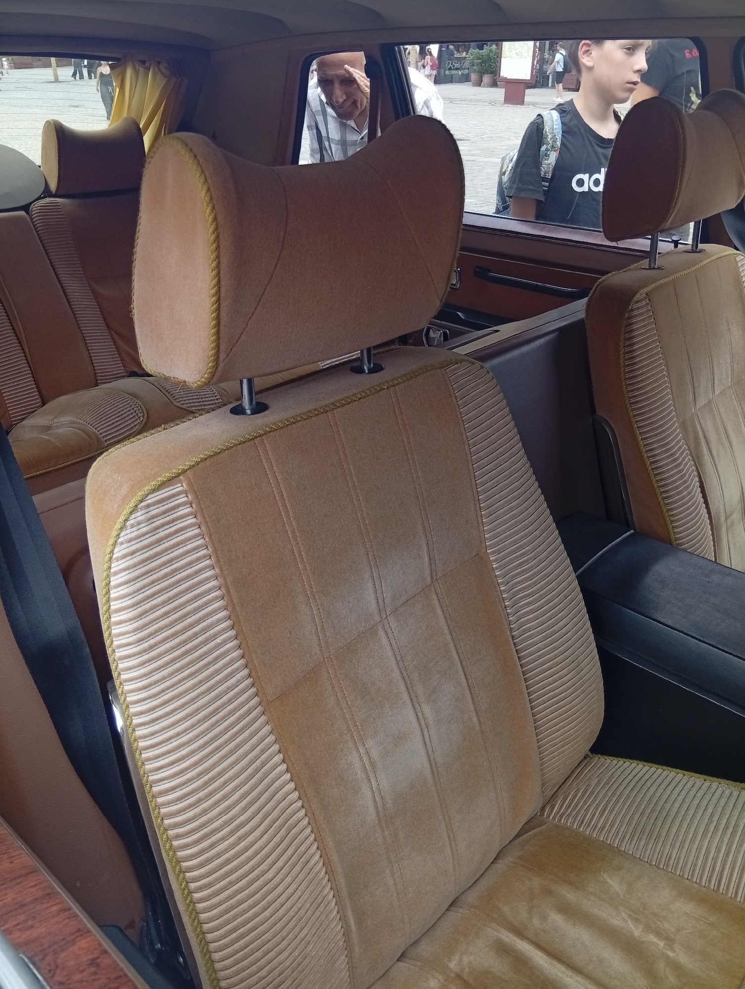 The photograph displays the interior of a vintage vehicle, focusing on the brown velour seats and headrests. The seats have a textured pattern on the sides and are trimmed with a decorative cord. Through the vehicle's windows, glimpses of an outdoor urban setting are visible. In the background, to the left, a man in a light-colored checkered shirt is looking into the vehicle. To the right, a teenage boy wearing a black t-shirt and a backpack is visible outside the vehicle. The environment outside suggests a public space, possibly a square or street, with some buildings and people in the distance. The overall impression is that of a paused moment, capturing the interior of a classic car with activity occurring outside. The image was taken in Wrocław, Poland.