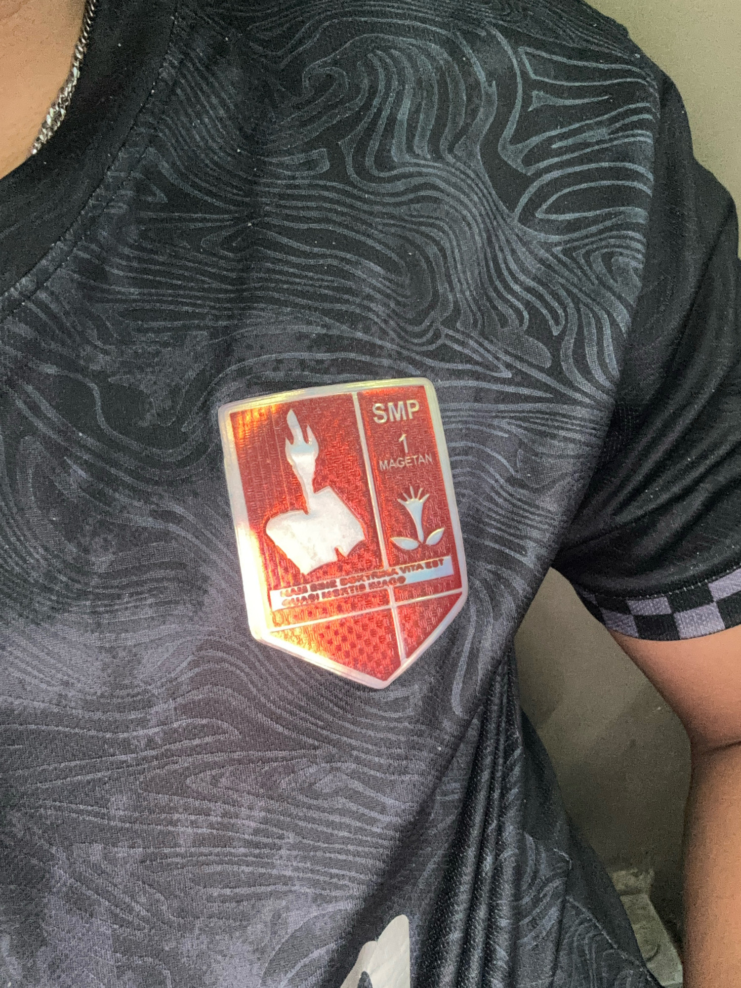 A person's chest is shown, wearing a black t-shirt with a repeating wavy pattern. A crest, approximately 3 inches tall and 2 inches wide, is attached to the left side of the t-shirt. The crest is shield-shaped with a red background and a metallic border. It contains a white graphic of a stylized torch and flower. Text within the crest reads "SMP 1 MAGETAN" at the top, and below it, "KARYA BHAKTI DOKTRIN BANGSA" and "JAYAKUSUMAH". The person is likely in an indoor setting, as indicated by the muted, solid background behind them.