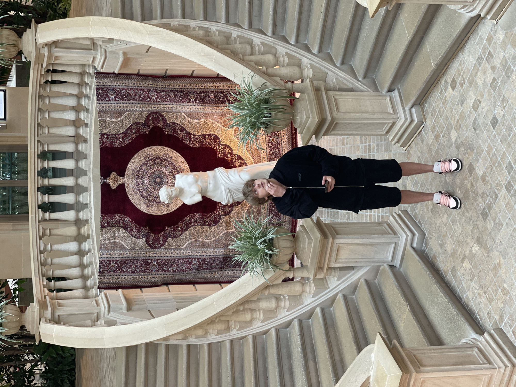 A young person stands in the center of a grand, indoor staircase area. They are looking upwards, with their right hand touching their chin, and holding a phone in their left. The staircase is made of stone with ornate balustrades and decorative urns holding plants on either side of the person. Behind them, a large, ornate rug with intricate patterns hangs on the wall, framing a white statue of a classical figure. The floor is tiled in a mosaic pattern. This scene is located in the city of Khimki, Russia.