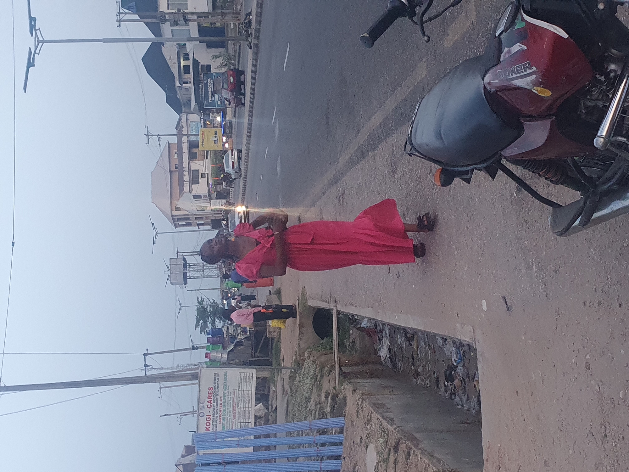 A scene on a street in Lokoja, Nigeria. A woman in a red dress stands on the sidewalk. A red and black motorcycle is parked nearby. Across the street, a sign for "Kogi Cares" is visible, and a "Thermocool" billboard. Cars and buildings line the opposite side of the street.