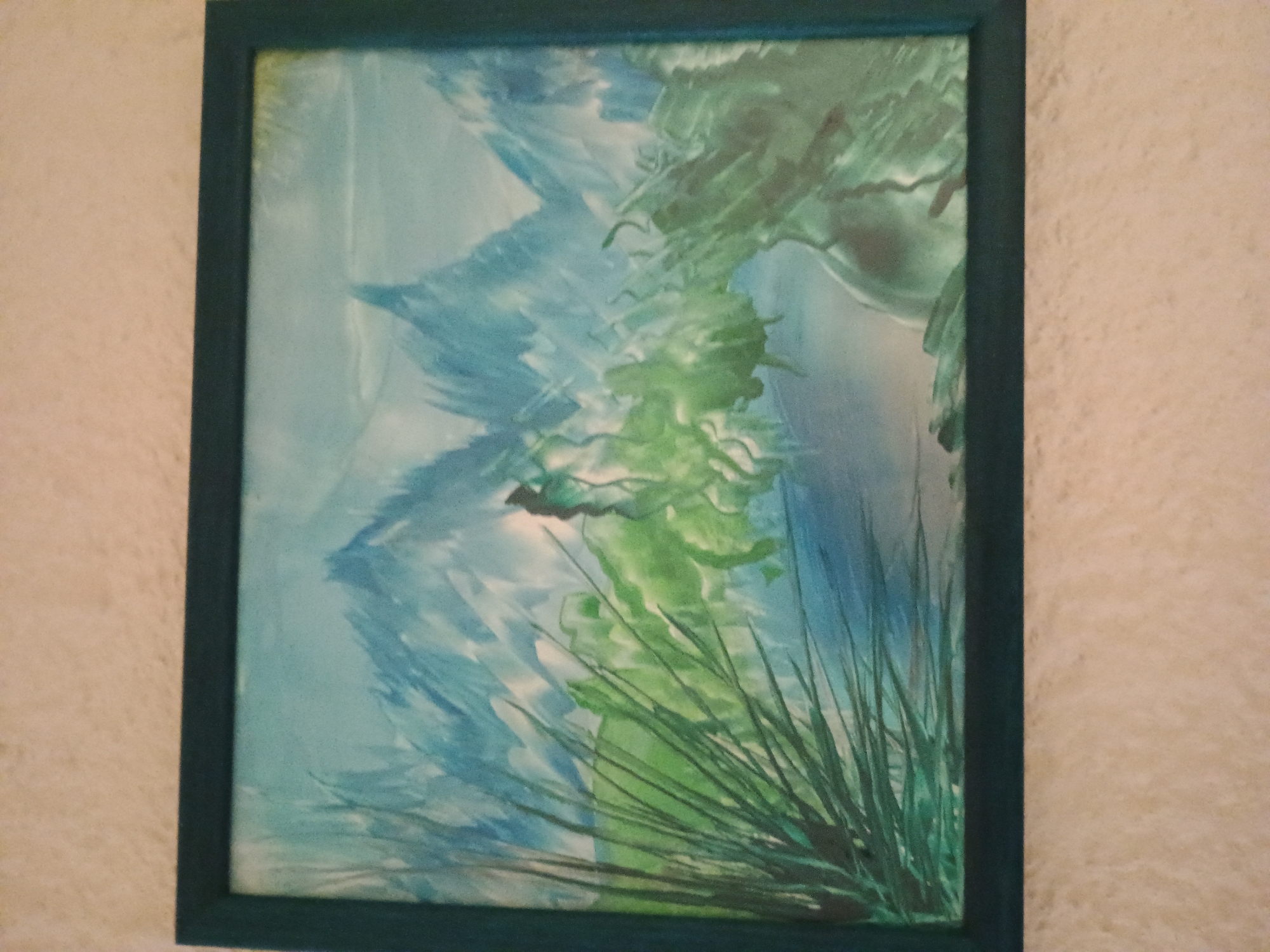 A framed abstract painting hangs on a beige wall. The painting is primarily blue and green, with strokes suggesting plant life or water. The frame is a dark green color. The painting is located in Unknown, Unknown.