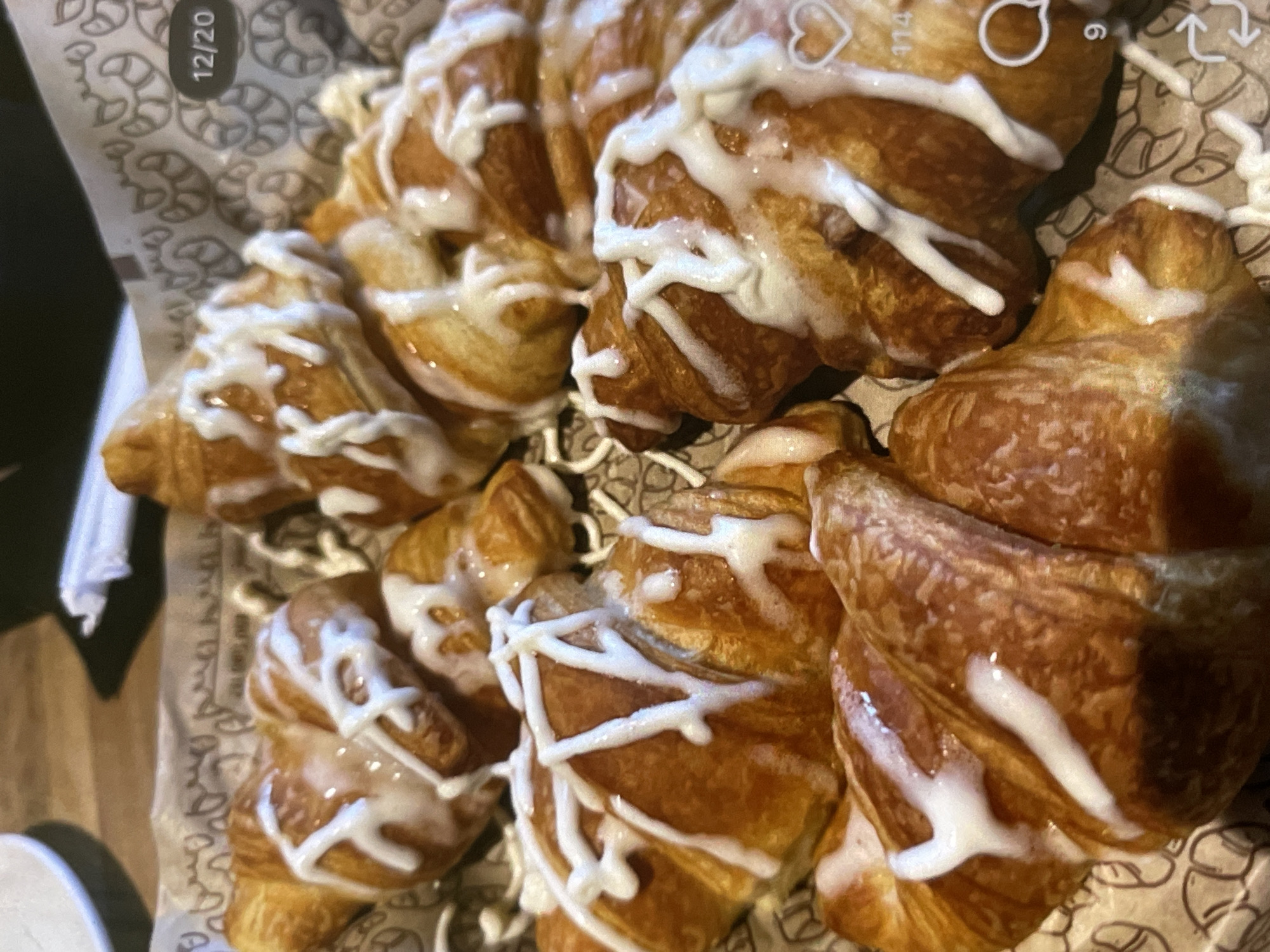 An overhead shot captures a close-up view of several glazed croissants arranged in a brown paper-lined basket. The croissants are golden brown with a flaky texture and are drizzled with white icing. The basket lining features a repeating pattern of croissant-like shapes. In the upper right corner, a "12/20" overlay is visible, suggesting a date or count. Other overlays include a heart icon and a speech bubble with the number "9" below it. The background is blurred, but appears to be a table with wooden texture. The scene is static and focuses solely on the pastries.