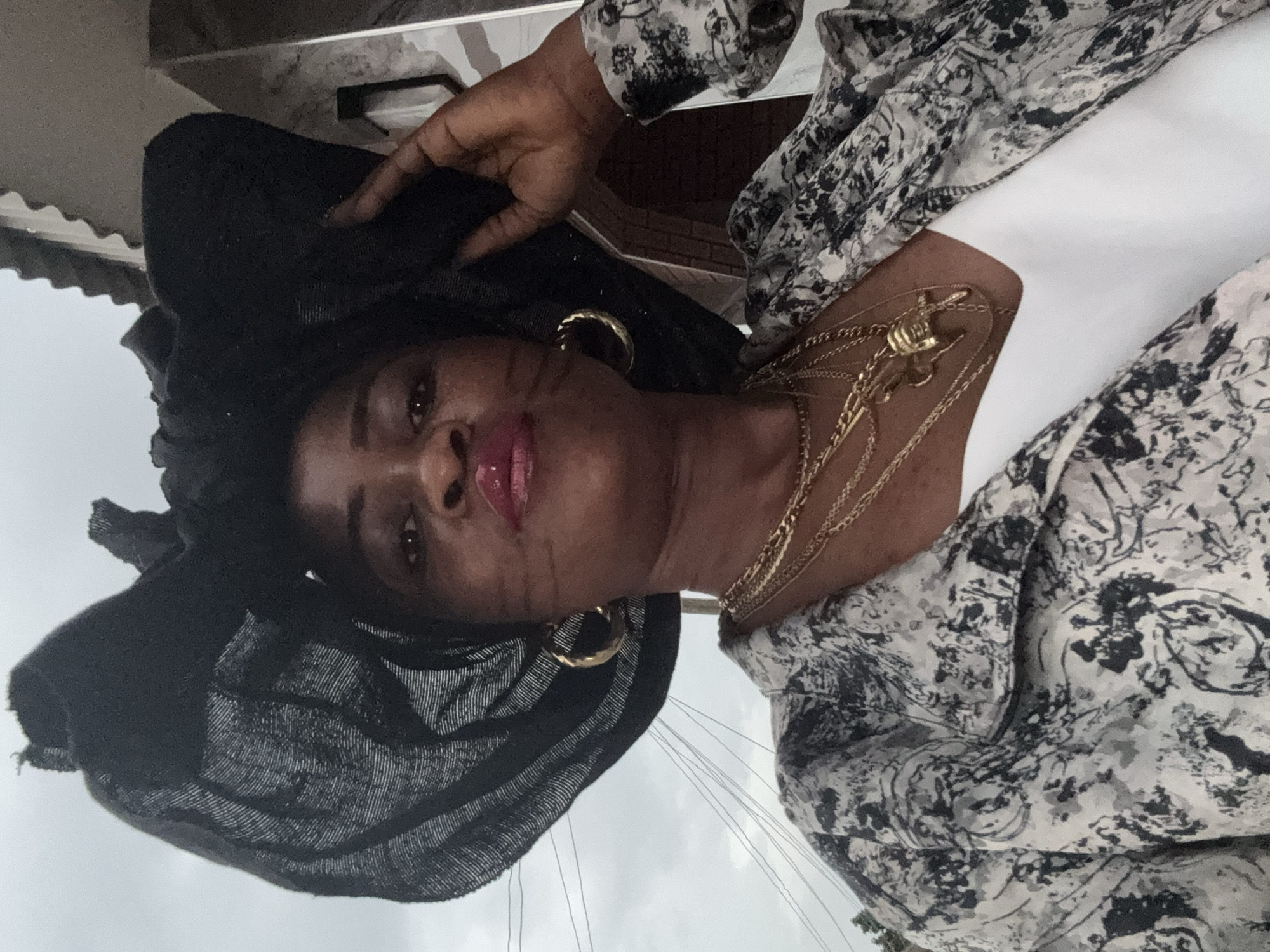 A woman with dark skin and dark hair is captured in a close-up, frontal shot, wearing a large, black, wide-brimmed hat adorned with fabric ruffles. She has on a white V-neck top and a black and white patterned jacket, layered with multiple gold necklaces, including one with a pendant. Large gold hoop earrings are visible, and her lips are painted with pink lipstick. Her right hand is raised, gently touching her hat. The background features an overcast sky with visible power lines and a portion of a building with a corrugated roof and brickwork, suggesting an outdoor setting in the city of Ajara, Nigeria. The overall impression is a formal portrait, possibly taken during an event or outing.