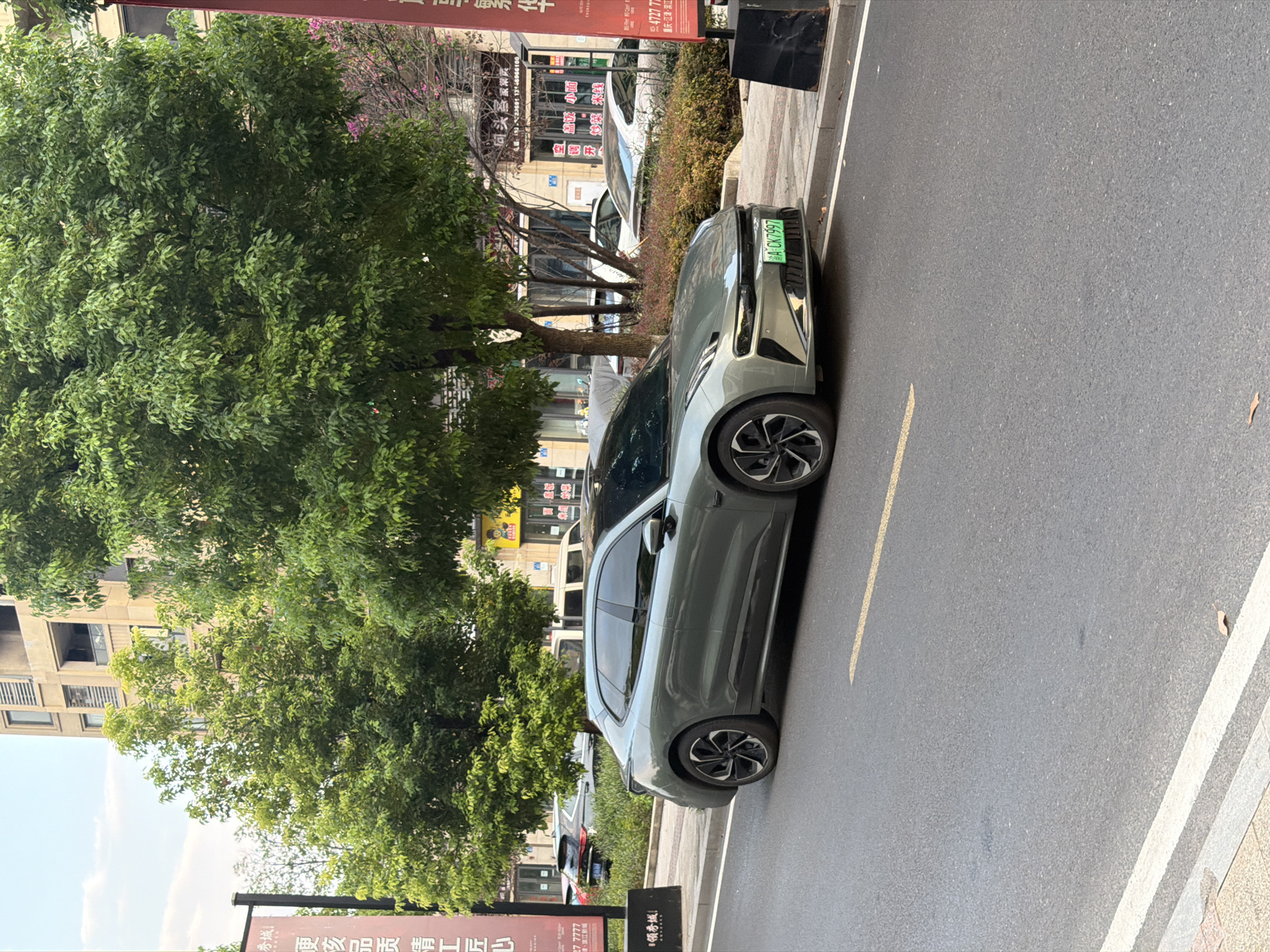 A light grey four-door sedan is parked on a city street in Jiangjin, China. The car is facing the viewer's left and is positioned at an angle, with its front end slightly closer to the camera. It has a sleek, modern design with a dark-colored grille and distinctive headlights. The license plate, visible on the front bumper, reads "A·CK7997". The car is parked on the right side of a paved road, marked with a yellow line.
To the right of the car, a black rectangular pillar stands near the curb. Behind the car and extending into the background are trees with dense green foliage. In the far background, a multi-story building with beige facades and windows is visible, along with other parked vehicles and storefronts. The scene appears to be an urban environment on a bright, clear day.
