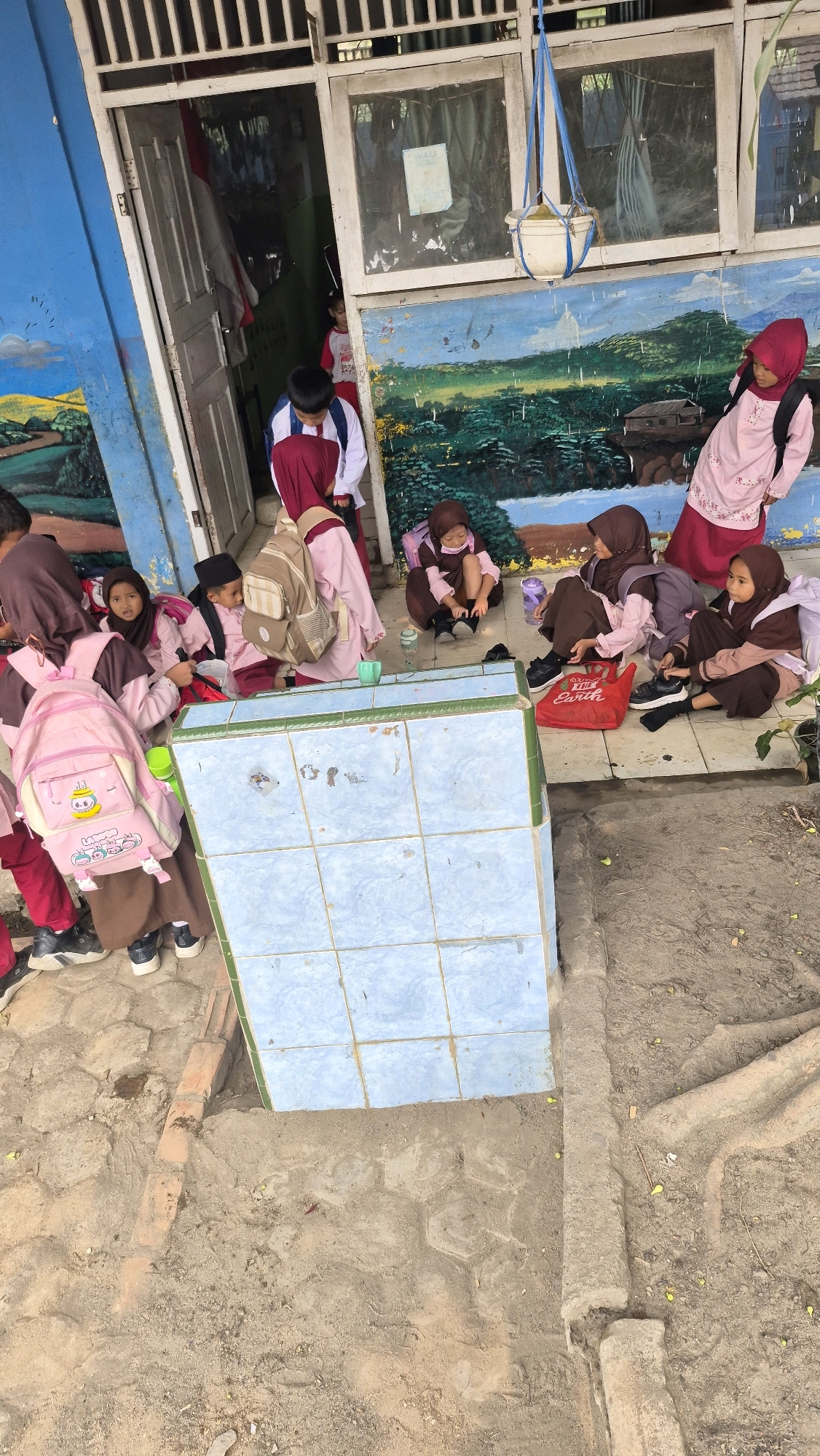 In an outdoor area near a building in Putak, Indonesia, a group of school-aged children, predominantly girls, are gathered. They are wearing uniforms that include pink hijabs and brown skirts or trousers, with some also wearing pink shirts. Several children are seated on the ground, some are adjusting their shoes, and others are interacting with each other. Backpacks, including a pink one with cartoon characters, are visible near the children. The environment includes a blue-tiled structure, a painted mural on the building depicting a landscape, and a dirt ground with some vegetation and brickwork. The scene appears to capture a moment of rest or transition for the students.