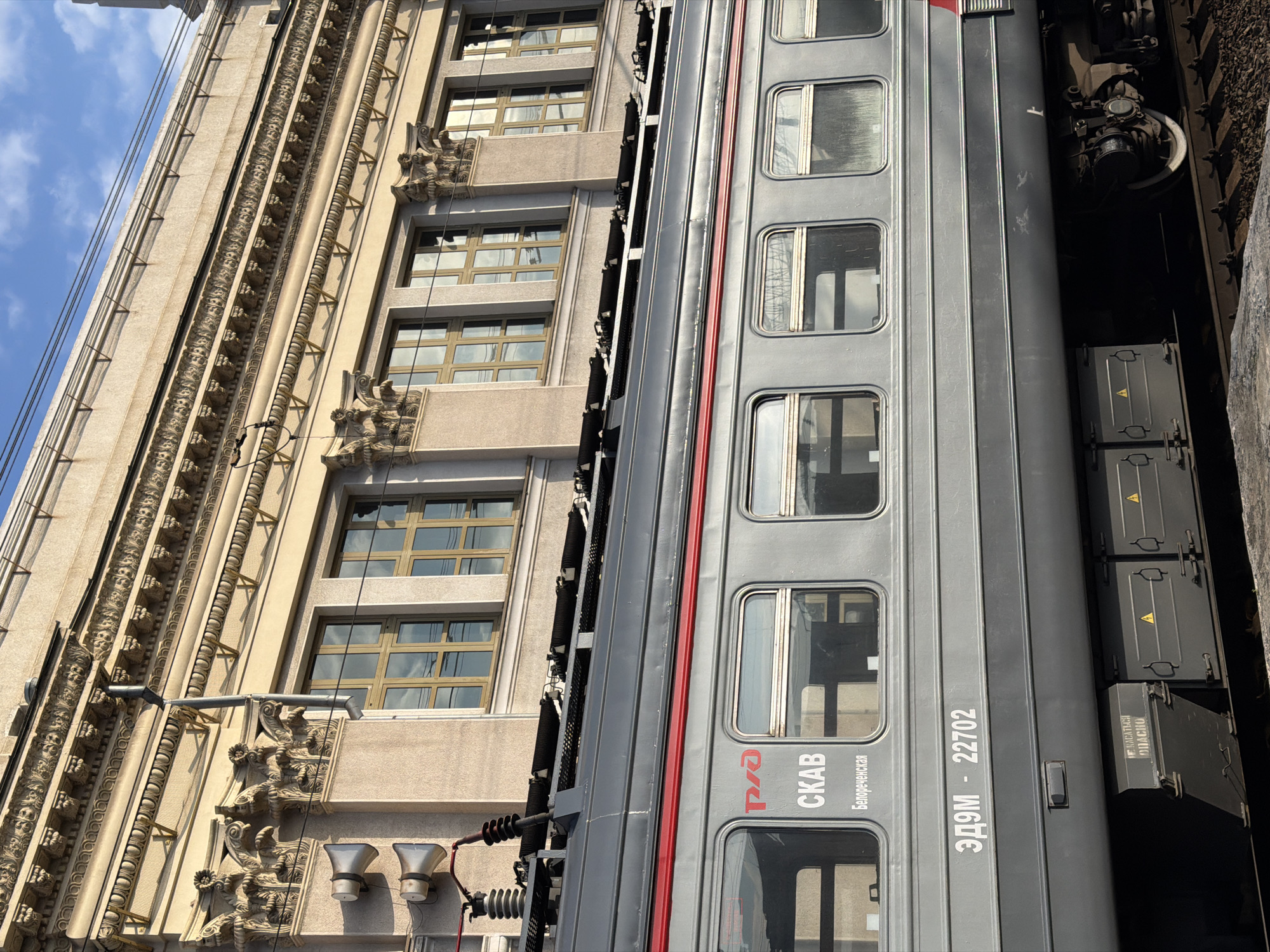A gray passenger train car, labeled "ЭД9М - 22702 СКАВ Белореченская" (ED9M - 22702 SKAV Belorechenskaya), is positioned next to a multi-story building with ornate architectural details. The building has multiple rows of windows. The sky is visible in the upper left corner. The train is on a track. The location is Krasnodar, Russia.