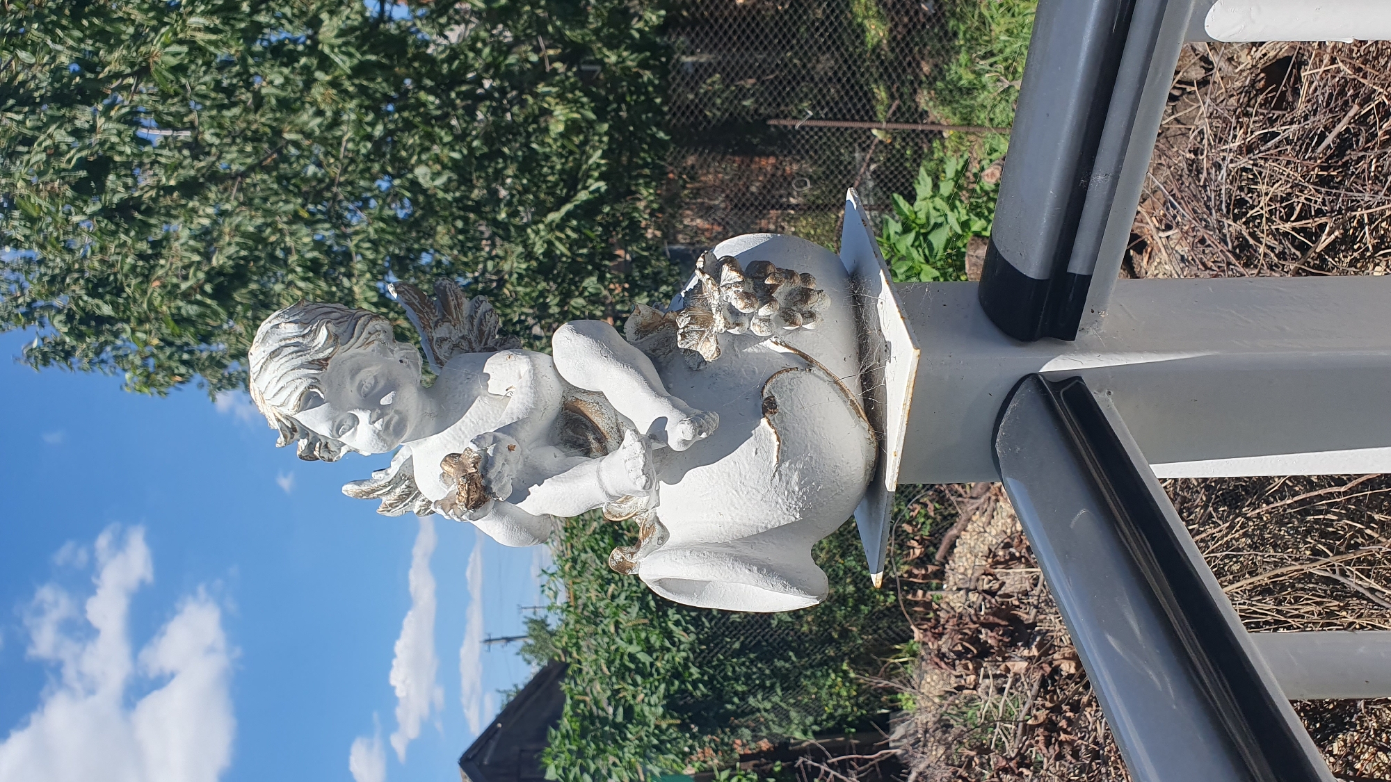 A white statue of a cherub holding a flower sits on top of a white pedestal. The cherub is depicted as a young child with wings and golden accents. The statue is outdoors, with a clear blue sky and some clouds visible in the background. Green trees and foliage are also present, along with a fence. In the foreground, a white railing with black accents runs horizontally across the frame. The scene appears to be in a backyard or garden setting in Ust-Labinsk, Russia.