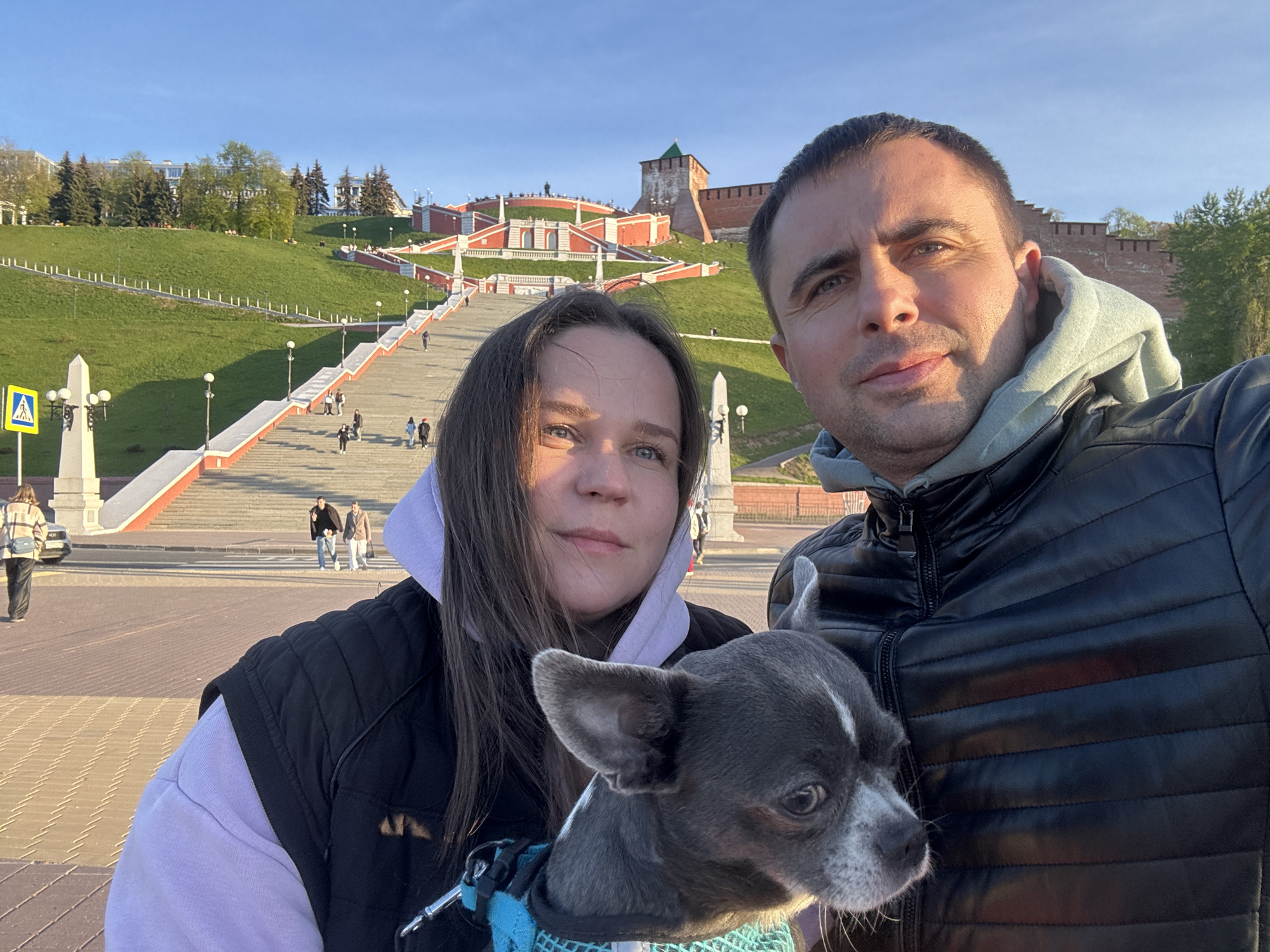 A daytime selfie features a man, a woman, and a small dog. The man is on the right, wearing a black leather jacket over a gray hoodie. The woman is on the left, wearing a black vest over a light purple hoodie. She is holding the small, gray and white dog with a turquoise harness. The background shows the Chkalov Staircase and the Nizhny Novgorod Kremlin in Nizhny Novgorod, Russia. People are visible walking on the staircase. There's a crosswalk sign on the left edge of the image.