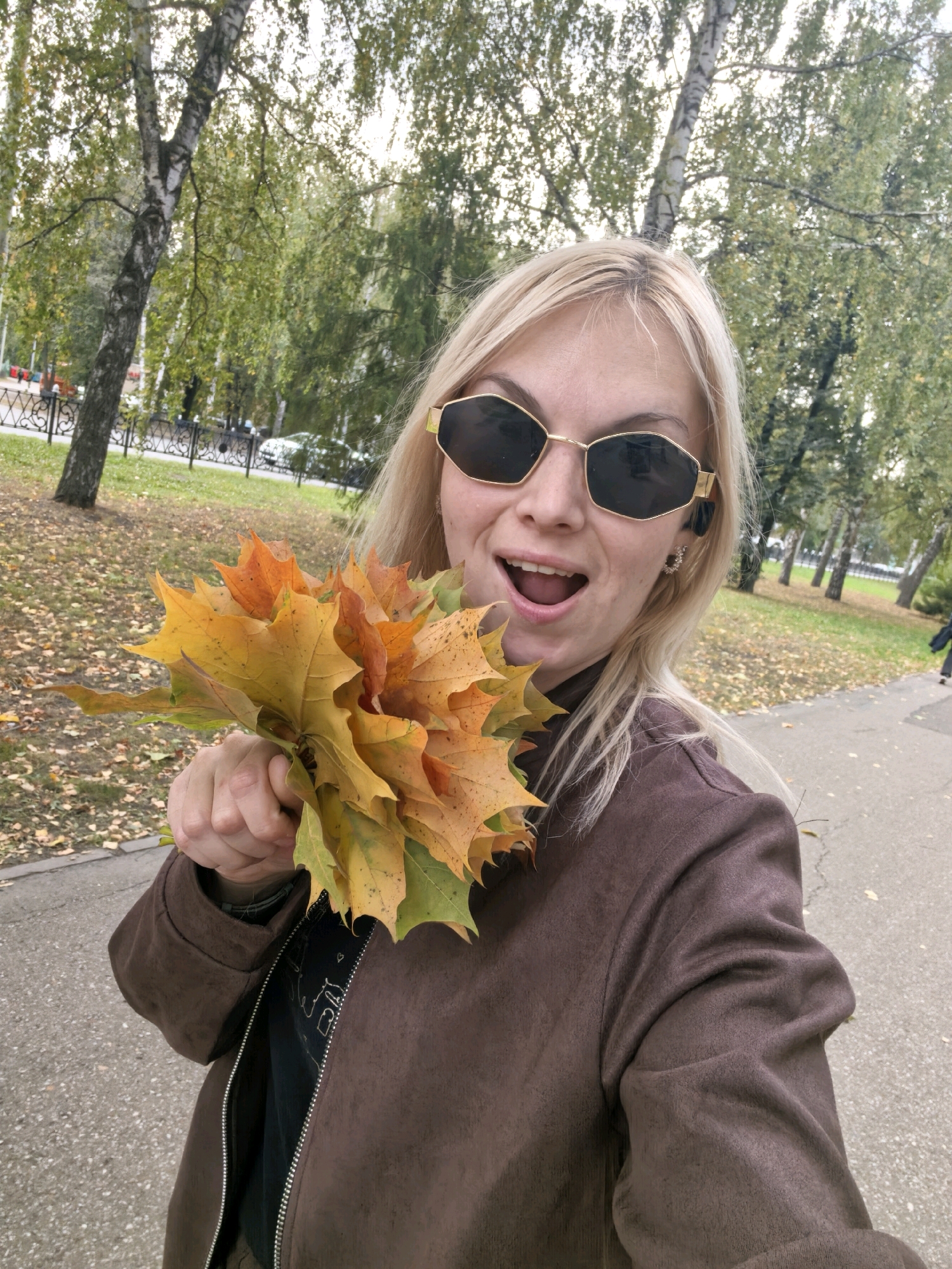 In Sterlitamak, Russia, a woman with blonde hair and sunglasses is taking a selfie in an outdoor park. She is holding a bouquet of autumn leaves in her right hand, which are a mix of yellow, orange, and green. She is wearing a brown jacket over a black shirt. In the background, there are trees with green and yellow leaves, a paved path, and a grassy area with fallen leaves. The sky appears overcast.