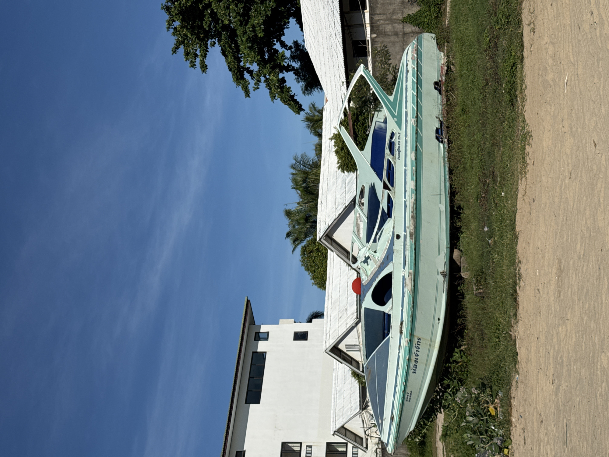 An abandoned, light green speedboat sits on dry land in front of a white building with a gray roof, with a clear blue sky overhead. The boat has a blue interior and the words "นอจเจ้าจักร" and "0047 50296" are visible on its side, along with Thai text that reads "กานเจา 25-2". To the right of the building, a large tree with dense green foliage is visible. The ground in front of the boat is covered with patchy green grass and some dirt. The scene appears to be located in a tropical or subtropical region, given the type of vegetation.
