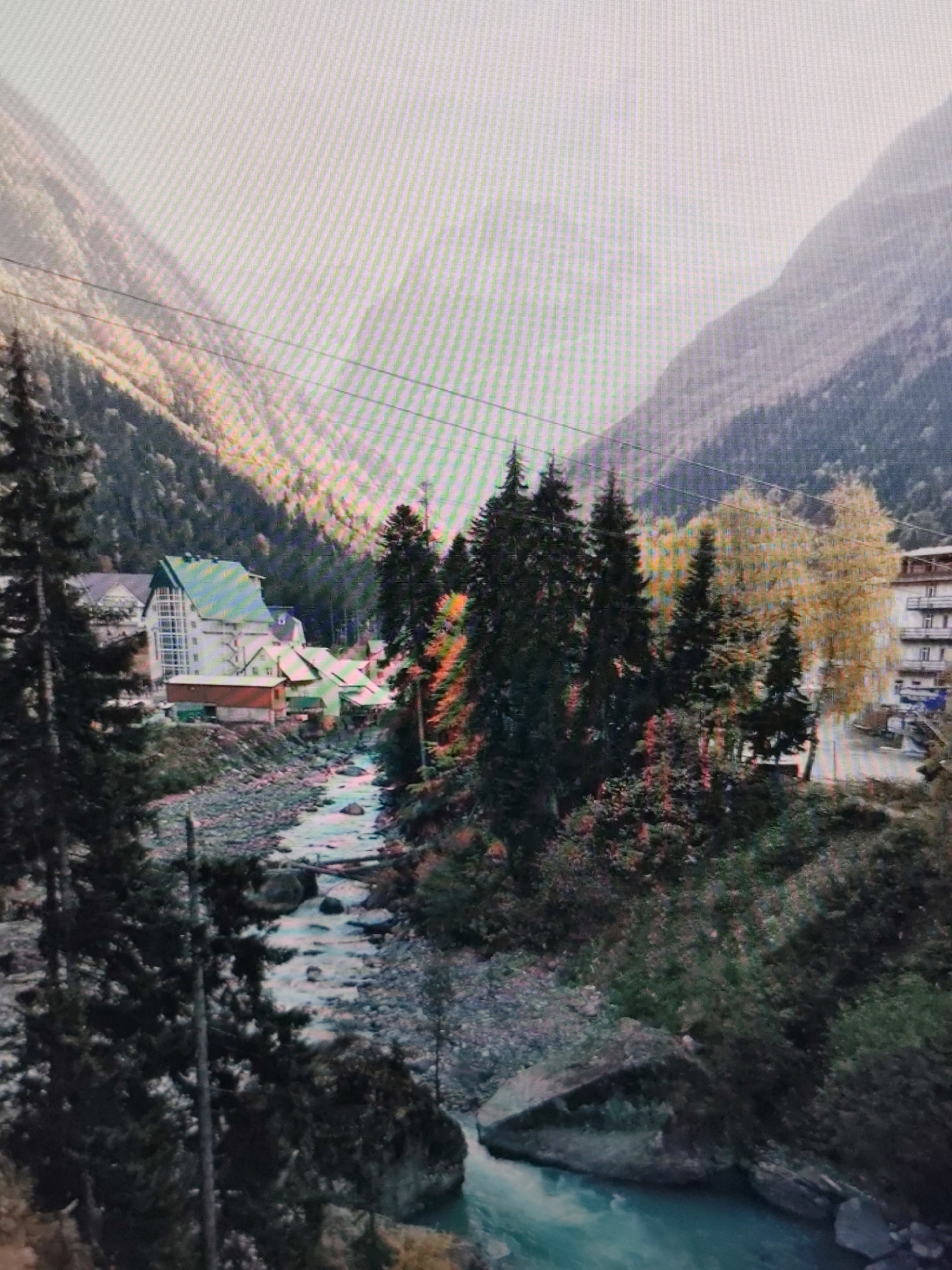 The image depicts a mountainous landscape with a river flowing through it. On the left side of the river, there are several buildings, including a prominent white building with a green, angled roof. Pine trees and deciduous trees with yellow and red foliage are scattered throughout the scene, suggesting an autumn setting. The river is rocky and turbulent, with visible logs or debris in the water. The mountains in the background are covered in dark green trees, with the sky being a bright, hazy white. There are no people visible in the image. The scene appears to be a rural or mountainous village setting, not a city like Syktyvkar.