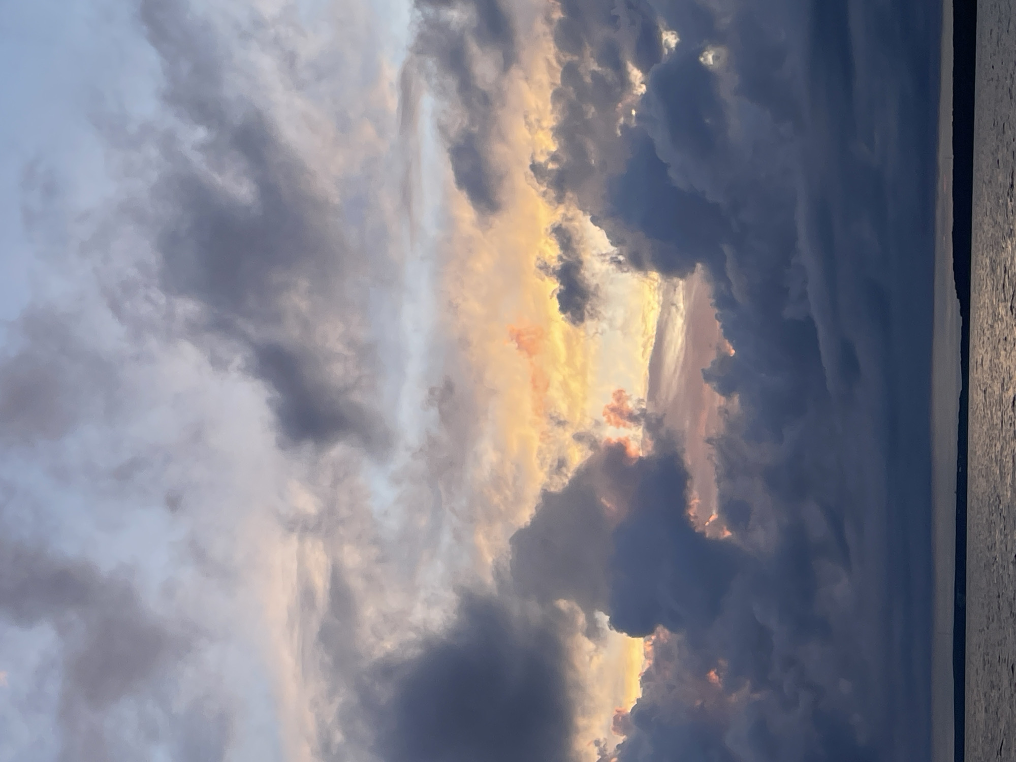 The image depicts a dramatic sky filled with dark, heavy clouds at sunset. Streaks of pink, orange, and yellow light pierce through breaks in the clouds, creating a striking contrast. The lower portion of the image shows a calm body of water, likely a lake or sea, with a distant, dark shoreline. The overall mood is atmospheric and moody, characteristic of a sunset over water. The location is identified as Yablonovka, Russia. There are no people, subjects, or specific objects visible in the image, nor any discernible actions or interactions. The environment is dominated by the natural elements of sky and water.