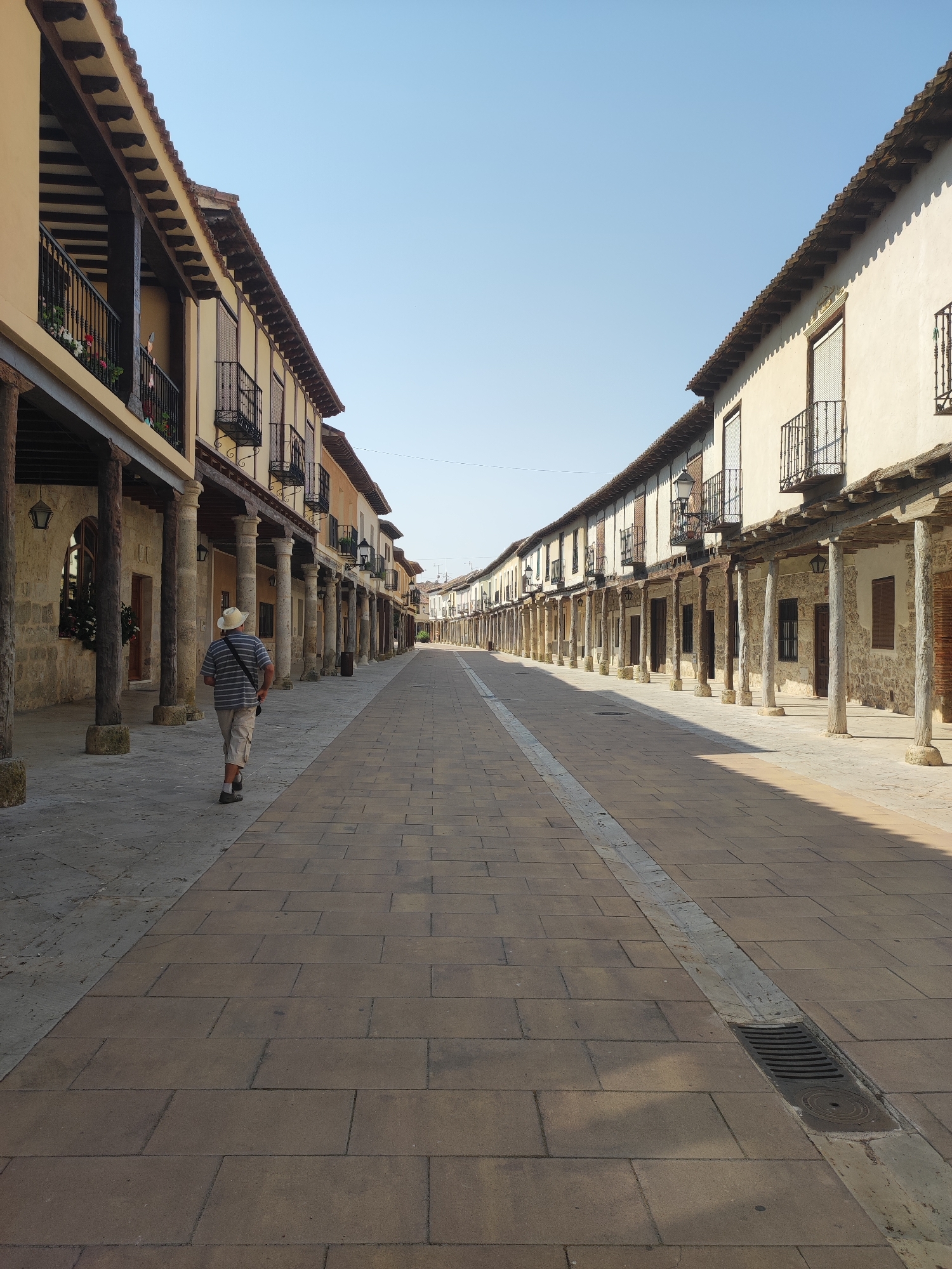 The image shows a man wearing a striped shirt, shorts, and a straw hat walking away from the camera down a long, paved street in Ampudia, Spain. The street is lined on both sides by traditional buildings with arcades supported by stone columns. The buildings have wooden balconies with wrought-iron railings and tiled roofs. The sky is clear and blue. The pavement is made of large, rectangular stone tiles, with a drainage grate visible in the center of the street. Sunlight casts shadows from the buildings and columns onto the pavement. The scene appears to be a historic part of the city, likely a plaza or a main street.