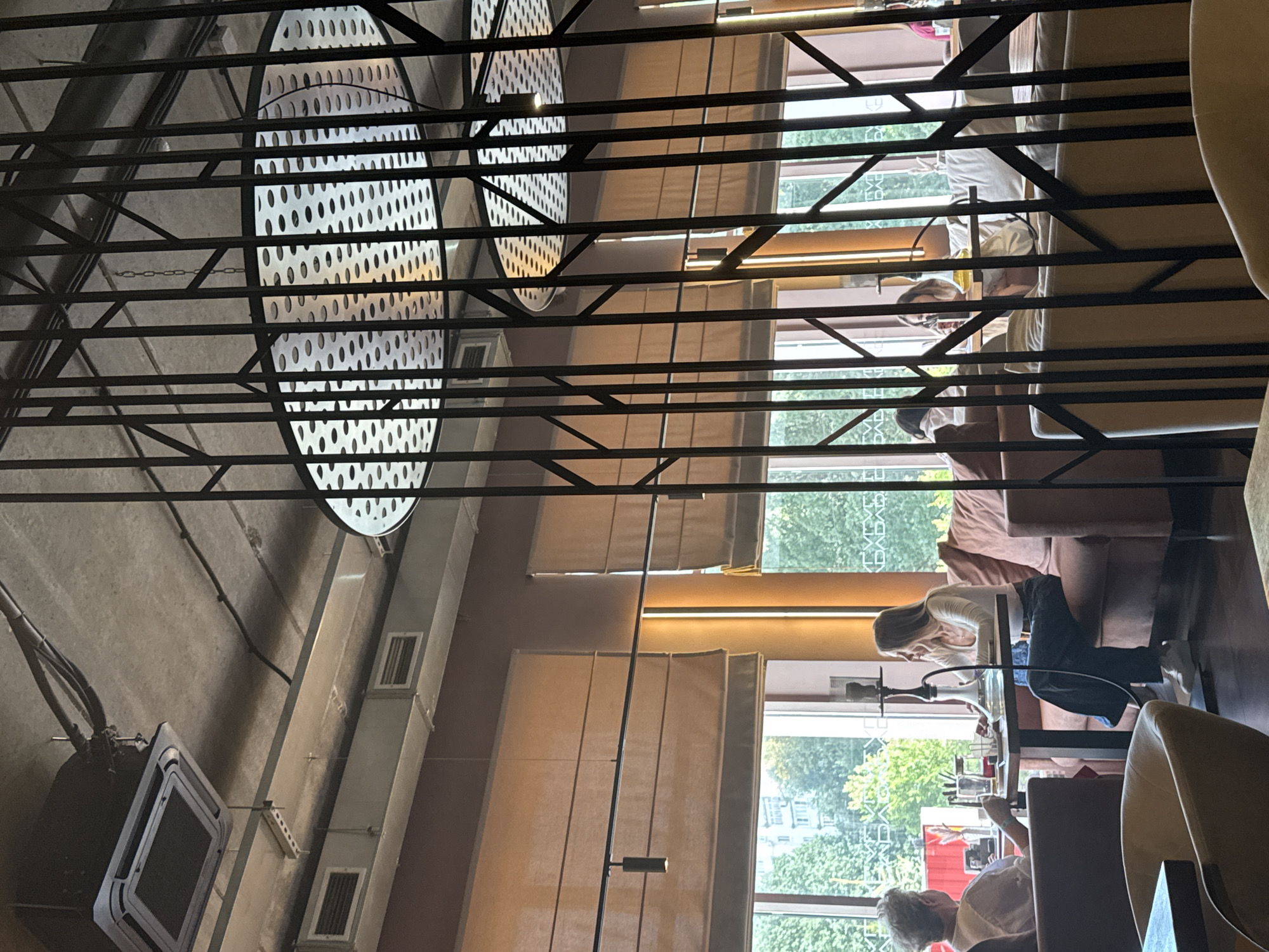 The image depicts the interior of a modern cafe, likely in Kazan, Russia, with elements of industrial and contemporary design. The ceiling is exposed concrete with visible ventilation systems and ductwork, characteristic of a loft or industrial-chic aesthetic. Suspended from the ceiling are large, circular light fixtures with perforated patterns, framed by an intricate black metal lattice structure.
In the foreground, seating areas are arranged with plush sofas and armchairs, some in neutral tones and others with pink upholstery. A low-lying table is partially visible in the bottom left corner.
Behind the lattice, a group of people are seated at tables near large windows that offer a view of lush green trees and buildings, suggesting an urban setting. Two women are the most prominent figures. One, seated on the left, has grey hair and is looking away from the camera, engaged in conversation or activity at her table. The other woman, in the center, is wearing a white top and dark pants, facing her companion and interacting with what appears to be a hookah setup on the table. Another person is partially visible behind her, and further back, two more individuals are seated. The windows are covered by light-colored Roman blinds, which are drawn down partially.
The overall ambiance is relaxed and social, with natural light filtering through the windows. The cafe's design incorporates both functional elements like ventilation and decorative features like the metalwork and lighting, creating a stylish and comfortable space.