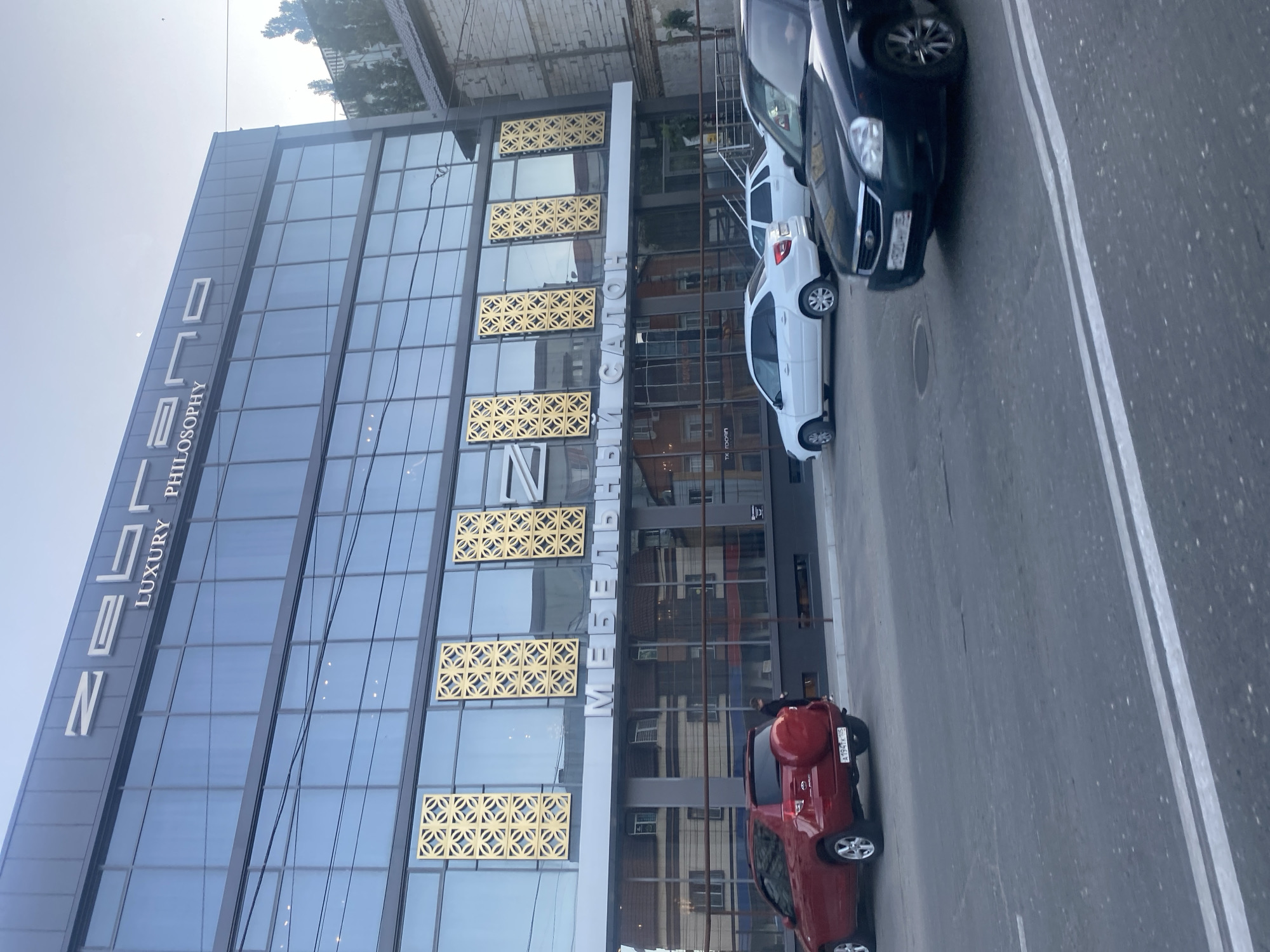 The image shows a modern building with a glass facade, featuring large windows and decorative gold panels. The building has prominent signage at the top, with the words "ZELENA LUXURY PHILOSOPHY" visible. Below this, another sign reads "МЕБЕЛЬ". Cars are parked along the street in front of the building, including a red SUV and several white and black cars. The scene appears to be in an urban setting, possibly a commercial area. The lighting suggests it is daytime, with the sky visible in the background. The overall impression is one of a contemporary commercial or residential complex in a city.