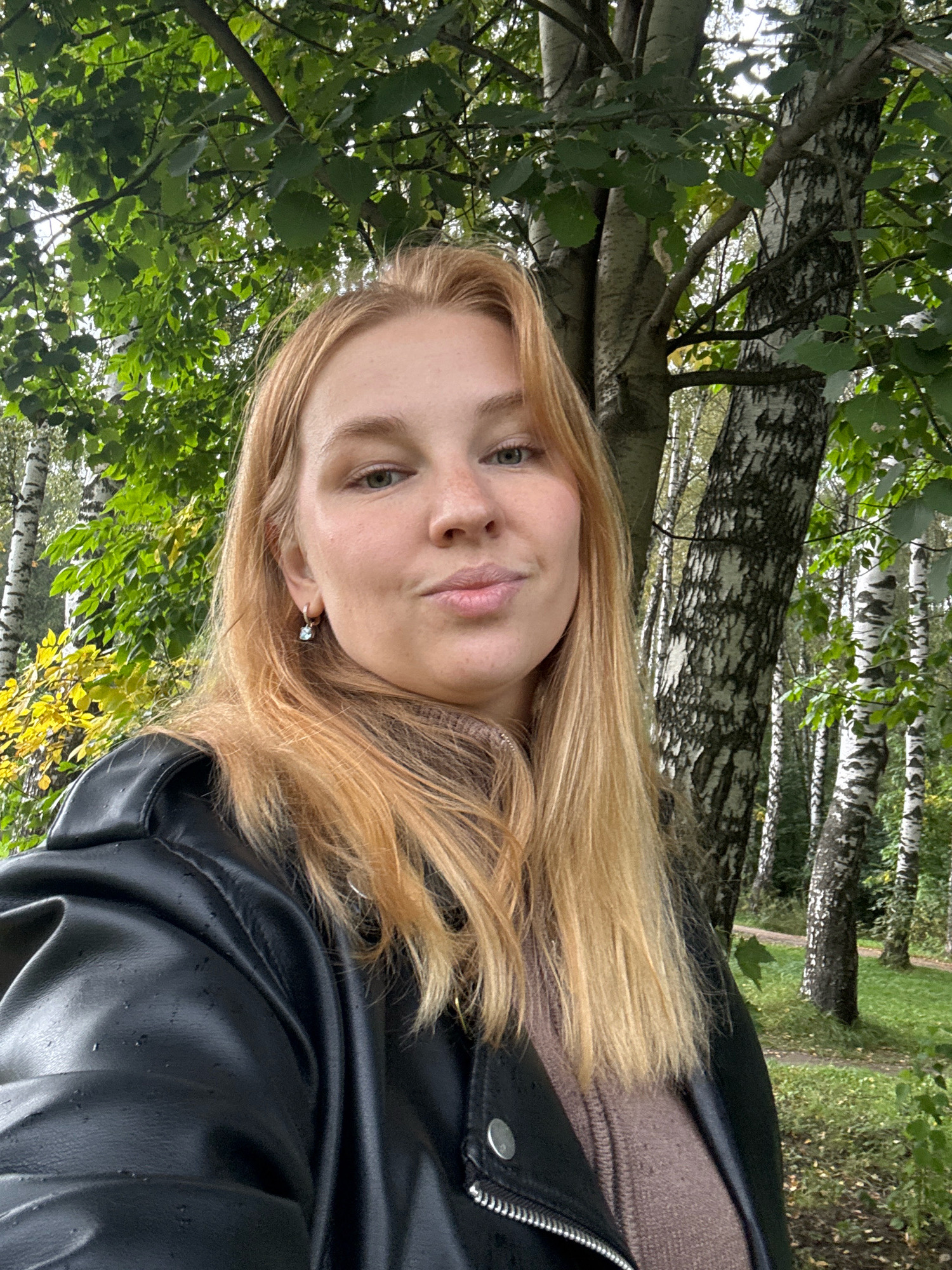 A young woman with blonde hair and fair skin is standing in a park, taking a selfie. She is wearing a black leather jacket and a brown sweater. The background shows a forest with birch trees and green foliage, suggesting an outdoor location in a city. The lighting is natural and appears to be overcast. The woman's expression is neutral, with her lips slightly pursed. The image is shot from a slightly low angle, emphasizing the trees above. The presence of birch trees and the general landscape are consistent with the city of Yaroslavl, Russia, which is known for its numerous parks and natural green spaces.