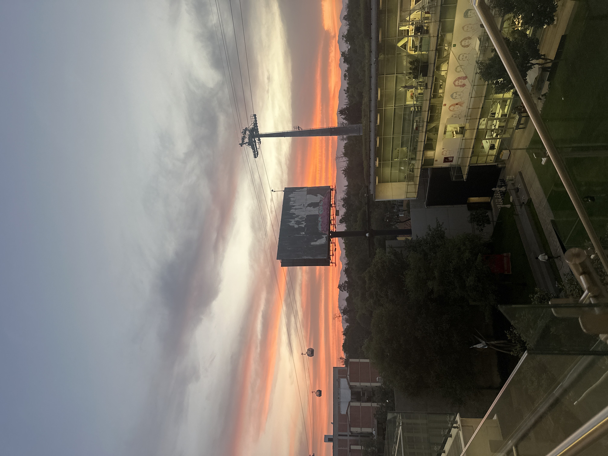 This image captures an urban landscape in Mexico City, Mexico, at sunset. The sky is a dominant feature, displaying a dramatic palette of vibrant orange, pink, and red hues transitioning into lighter greys and blues, indicating the setting sun. Scattered clouds are also visible, some reflecting the sunset colors.
A cable car system is prominent on the left, featuring a tall, concrete support tower and several modern gondola cabins suspended on overhead lines, moving across the frame. In the mid-ground, a large, rectangular billboard with faded or graffiti-covered advertising stands below the cable car lines. Beyond these structures, a silhouette of a mountain range is visible against the intensely colored horizon.
To the right, a series of modern, glass-fronted buildings rise into the frame. Many windows show interior lights turned on, suggesting offices or residential spaces. One particular building displays a large mural or print of stylized faces along its exterior. Below these buildings, there are green spaces, trees, and outdoor areas with seating. The foreground shows a glass railing, indicating the photo was taken from an elevated vantage point, such as a balcony or upper floor.
The scene depicts the transition from day to evening in a bustling urban environment, with the cable cars actively traversing their lines. No explicit readable text is visible, beyond the abstract nature of the mural and the graffiti on the billboard.