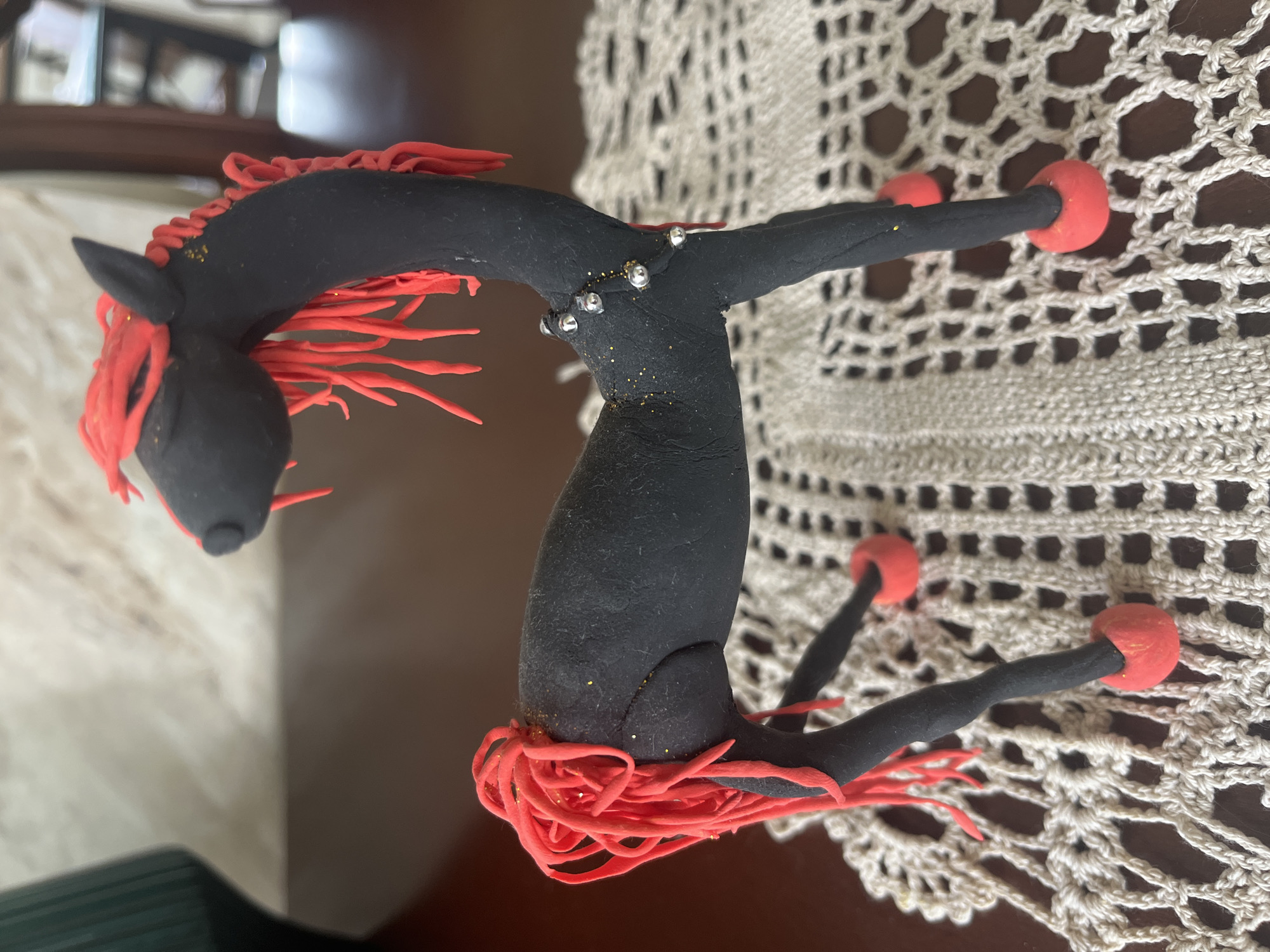 A black clay horse figurine with red yarn for its mane and tail stands on a crocheted placemat. The horse has red hooves and is decorated with small silver beads and gold glitter on its neck and hindquarters. The background is blurred, but appears to be an indoor setting with a wooden table. There is no indication of the location being in Vladimirovka, Russia.