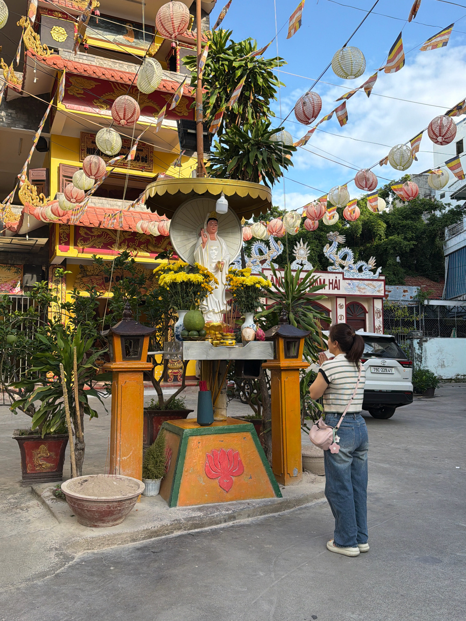 The image depicts a woman praying in front of a statue of a deity at a Buddhist temple in Nha Trang, Vietnam. The woman is wearing a striped t-shirt, blue jeans, and a pink crossbody bag. She is standing with her hands clasped in front of her chest, facing the statue. The statue is white and adorned with yellow flowers. The temple itself is colorful, with red and yellow accents and ornate decorations. The area is decorated with numerous paper lanterns and prayer flags, suggesting a festive or religious occasion. In the background, a white SUV is parked, and lush greenery is visible. The ground is paved with concrete.