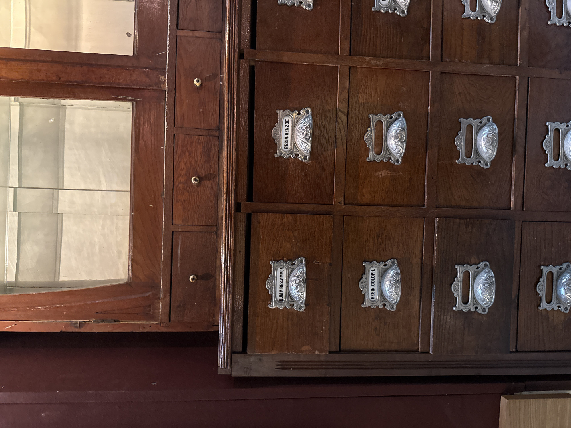 The image depicts a portion of an antique wooden cabinet with glass-fronted display on the left, and multiple drawers on the right. The drawers have ornate, silver-colored handles with labels, including "RESIN BENZOE", "GUMMIRES. AMMON", and "RESIN. COLOPH.". The cabinet appears to be located in a historic pharmacy or apothecary, likely in Pazardzhik, Bulgaria. The environment is dimly lit, suggesting an indoor setting.