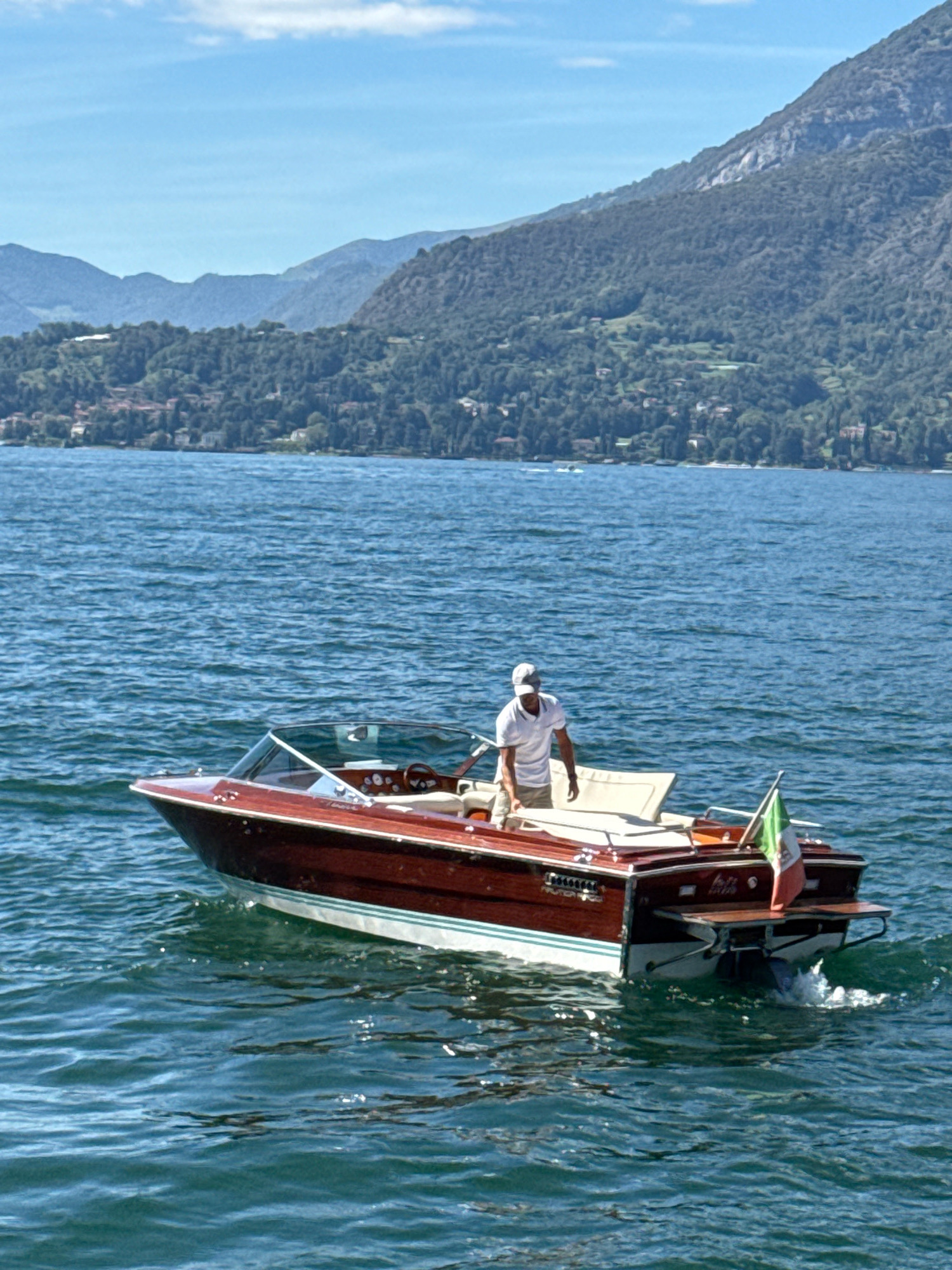 A wooden speedboat with a white interior is cruising on a blue body of water. A man in a white t-shirt and a baseball cap is standing at the helm of the boat, looking to the right. An Italian flag is attached to the stern of the boat. In the background, lush green hills dotted with buildings and trees rise towards a clear blue sky. The boat appears to be in motion, leaving a small wake behind it. The scene suggests a leisurely day on the water, possibly in the vicinity of Varenna, Italy, given the characteristic landscape of the Italian lakes.