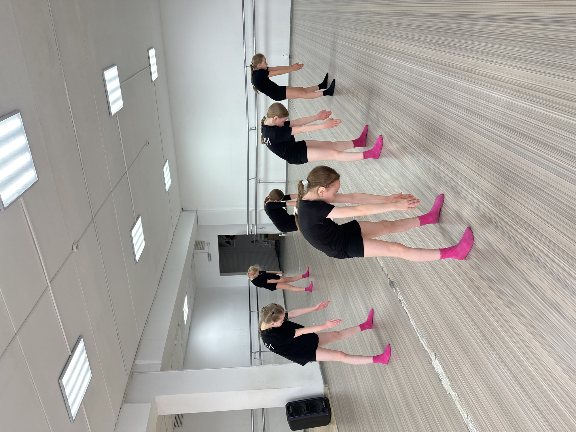 An image captures a group of young girls performing an exercise in what appears to be a dance or fitness studio. The main photo is rotated, showing the scene on an angle, but the actual orientation depicts the girls lying on their backs on a light-colored, striped wooden floor. Their heads are towards a white wall with ballet barres.
There are approximately six girls visible, though some are reflections in the large mirrors that line one wall. They are uniformly dressed in black t-shirts and black shorts. Most are wearing bright pink socks, while one girl at the back has black socks. Their hair is neatly pulled back, primarily in ponytails or braids. They are performing an exercise where they lie on their backs, extend their legs upwards with feet off the ground, and stretch their arms forward, parallel to their legs, with palms facing each other. Their expressions show concentration and focus.
The studio has a clean, minimalist aesthetic with white walls and ceiling. The ceiling features several rectangular fluorescent light panels, and a smoke detector is also visible. Along the back wall, a sign with the Russian word "ВЫХОД" (EXIT) is present, along with an air vent, confirming the location context of Votkinsk, Russia. A black portable speaker is placed on the floor in the foreground. The lighting is bright and even, typical for an indoor exercise space. The large mirrors reflect the room and the girls, creating a sense of depth and activity.