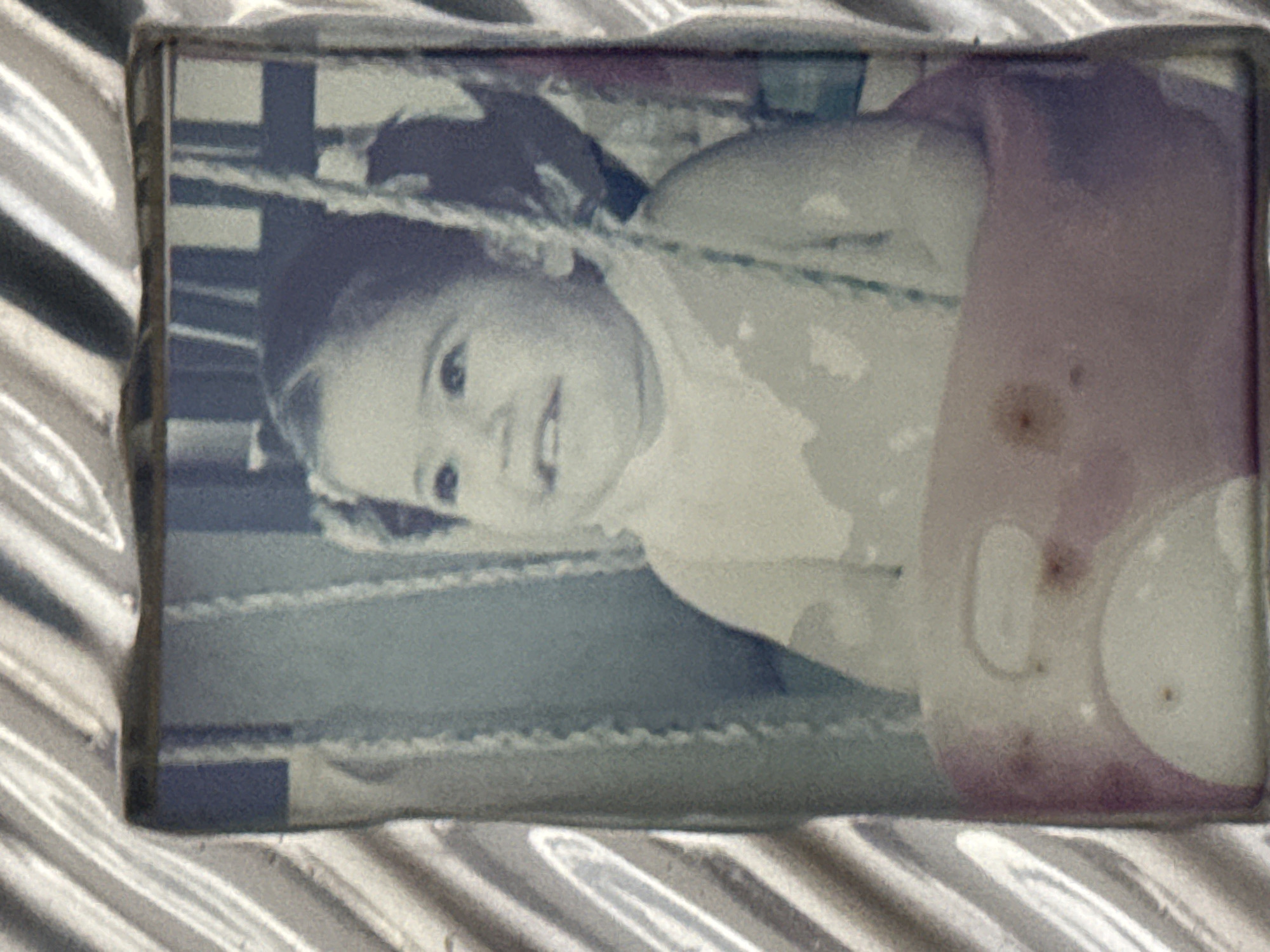 A framed photograph shows a young child, possibly a girl, facing the viewer with a slight smile. The child appears to be riding on a toy or swing, with a portion of a rounded object visible to the right of the child. The photo has color fading and imperfections, and is contained within a silver, patterned frame. It is not possible to determine the location in the city of Santa Clara Ocoyucan, Mexico from this image alone.