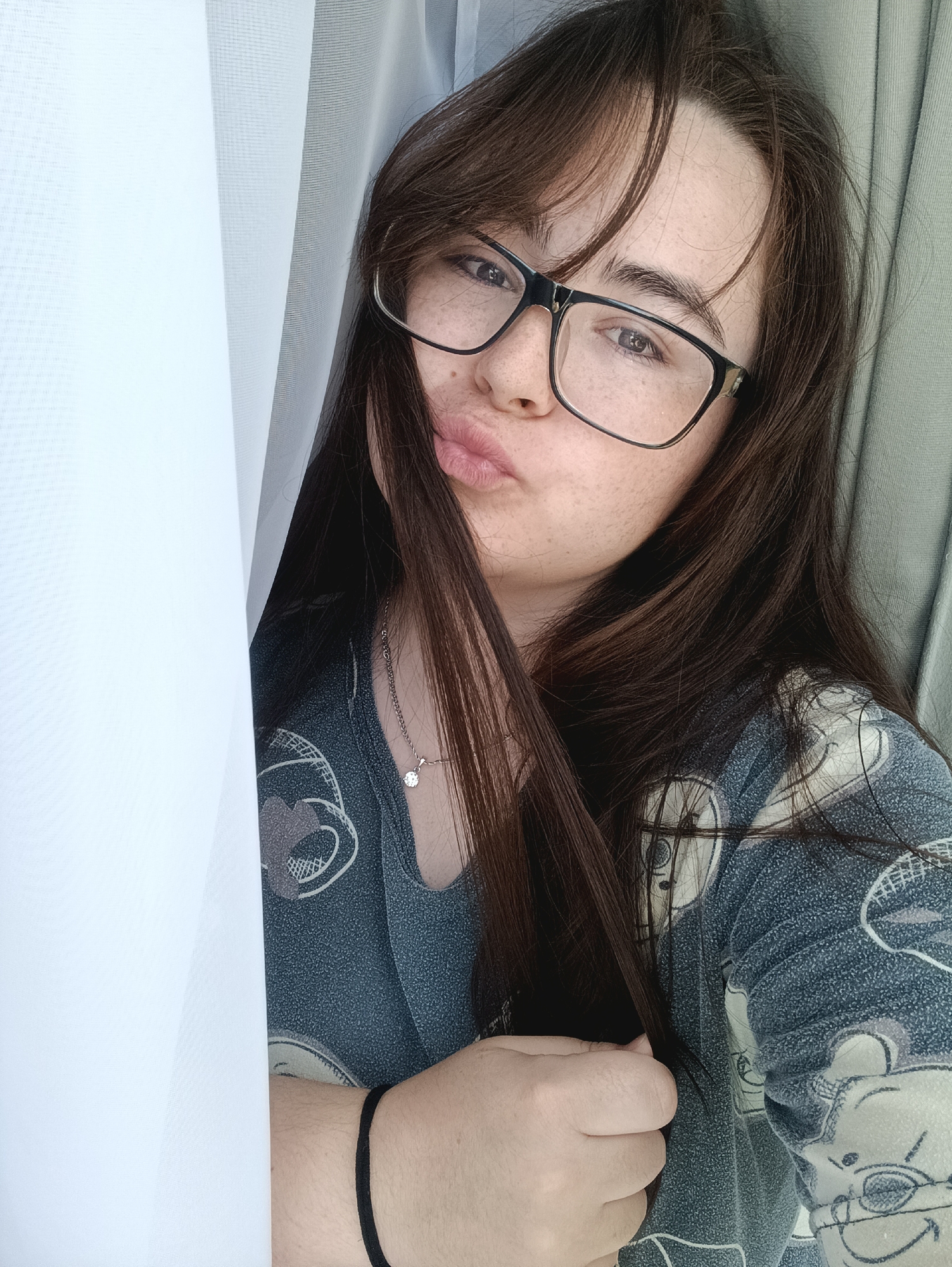 A young woman with dark hair and glasses is posing for a selfie in Noviy, Russia. She is wearing a gray pajama top with a cartoon character print. The background consists of white and gray curtains, suggesting she is indoors. She has freckles and is puckering her lips, with her hair partially obscuring her face. A silver necklace with a small pendant is visible on her neck, and a black bracelet is on her left wrist. The lighting is natural, coming from a window.