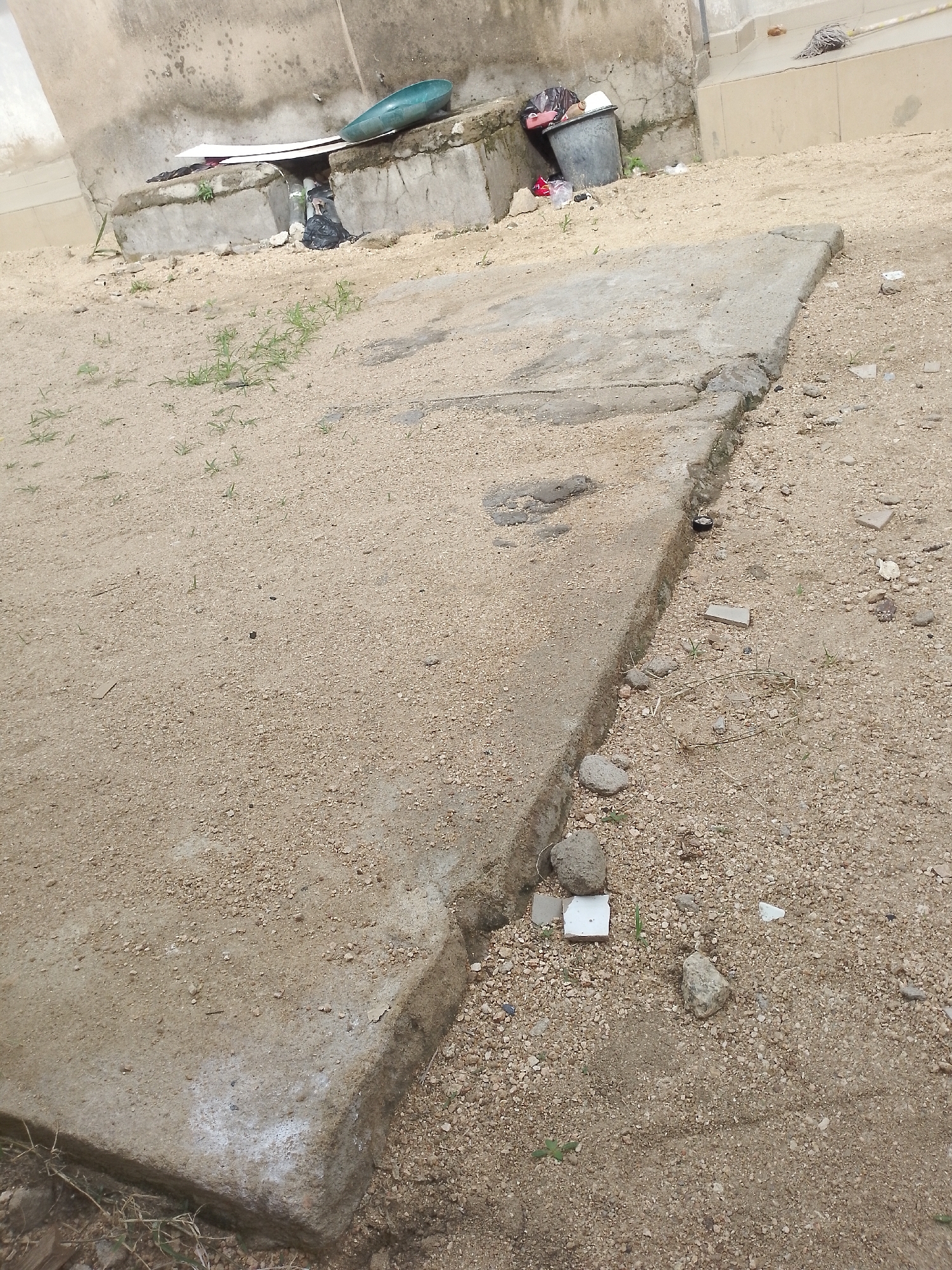 The image depicts an outdoor scene on a dirt and gravel surface in Jauro Yinu, Nigeria. A cracked concrete slab, which appears to be part of a pathway or foundation, dominates the foreground and extends diagonally across the frame. To the left of the slab, a wall made of concrete blocks is visible, with some debris and a blue basin resting on top. Further to the left, a grey trash bin and black garbage bag are present near the wall. The ground is covered with a mix of sand, small stones, and sparse green vegetation. The overall impression is one of a somewhat unkempt outdoor area.