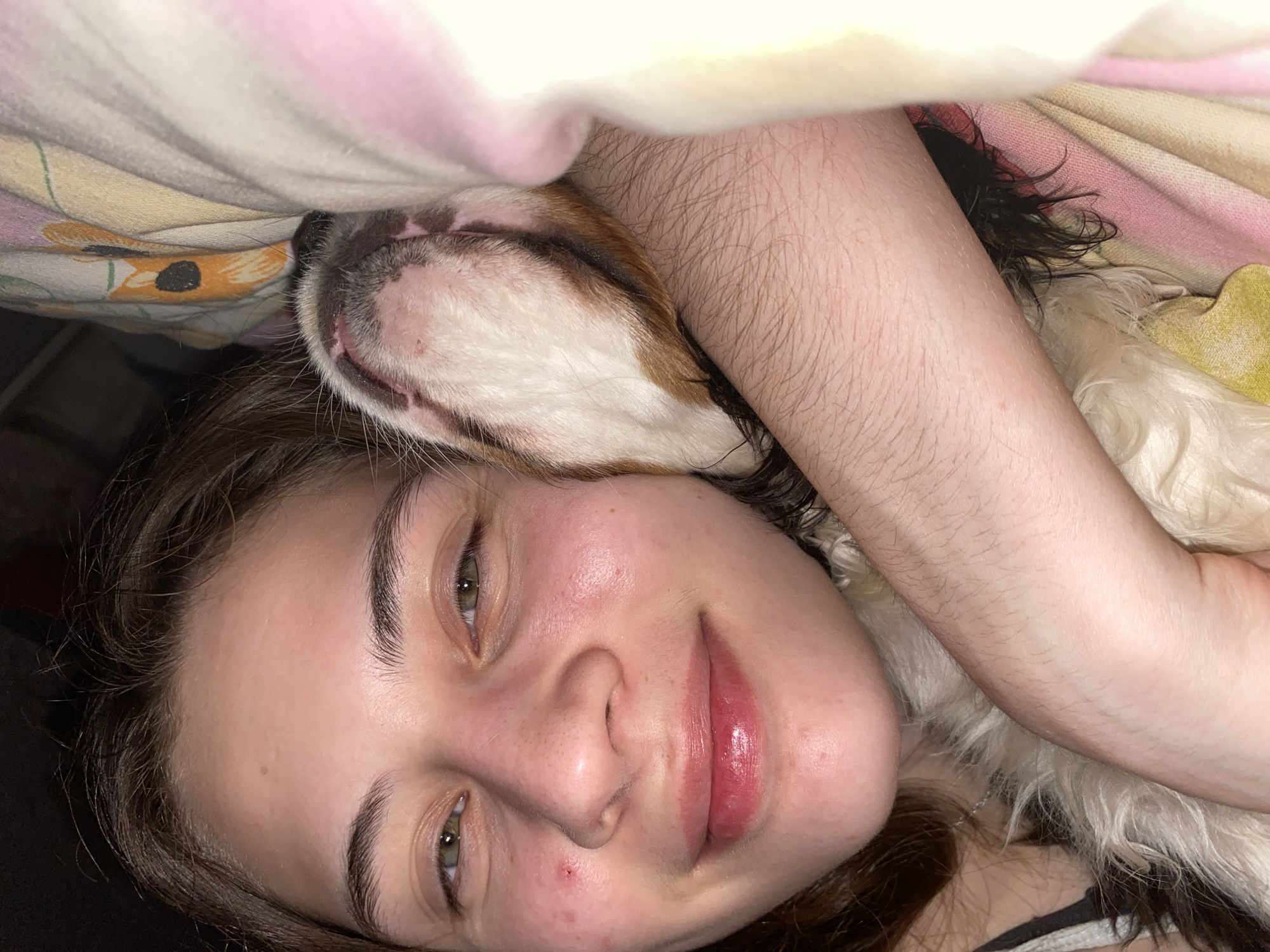 A young woman is lying down indoors, possibly in a bed, as indicated by the patterned bedding visible in the background. She is gazing at the camera and has a relaxed smile. A dog, primarily white with brown markings, is nestled close to her face. A person's arm is also visible, resting near the dog and the woman. The location is not specified or implied in the image.