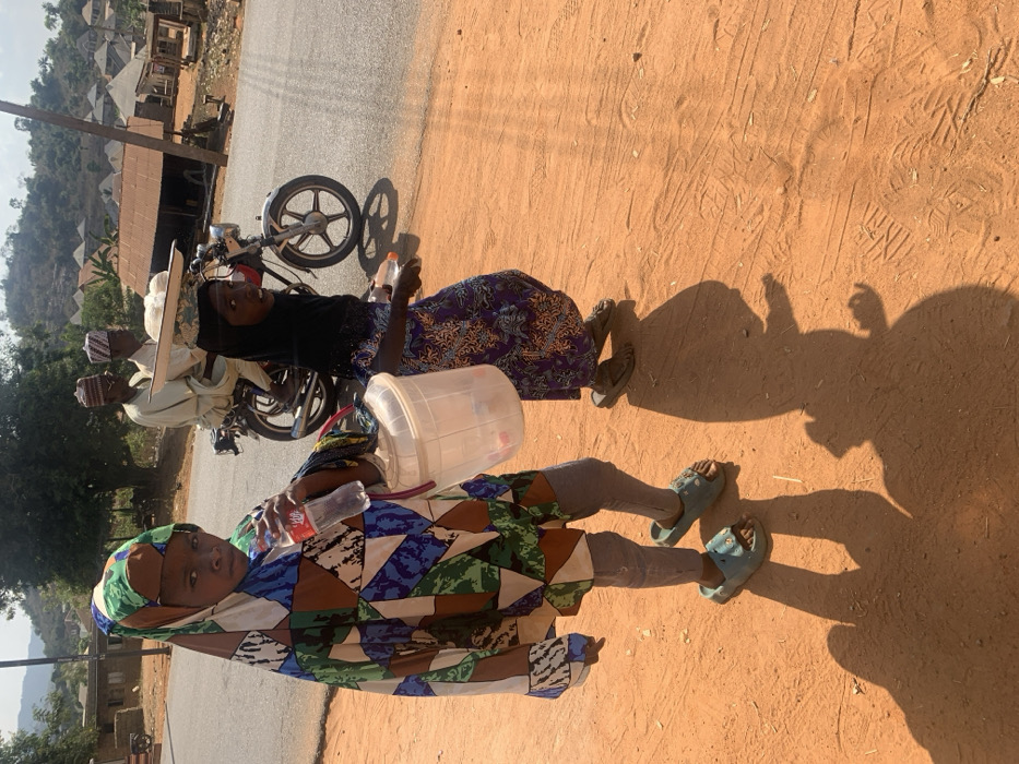 The image depicts a daytime scene in Egbedi, Nigeria, showing a partially unpaved environment adjacent to an asphalt road. In the foreground, two individuals, likely children, walk on a reddish-brown dirt path. The individual on the left, wearing a multi-colored patterned tunic and head covering, grey leggings, and light blue slides, holds a clear plastic Coca-Cola bottle in their right hand and a clear plastic lidded bucket in their left. This individual faces forward, making eye contact with the viewer. To their right, a second individual, wearing a dark head covering and a purple patterned dress with dark sandals, carries a small rectangular object in their right hand and walks towards the right of the frame, partially looking towards the viewer. Both individuals cast distinct shadows onto the dirt path. In the midground, on an asphalt road, a dark-colored motorcycle is in motion, carrying two adult males. Both males wear light-colored traditional attire and caps. The passenger, seated behind the driver, balances a flat, round tray containing several white, rounded objects on his head. The background features sparse green vegetation, hills, and a cluster of buildings with corrugated metal roofs, indicative of a rural settlement. The sky is bright and clear.